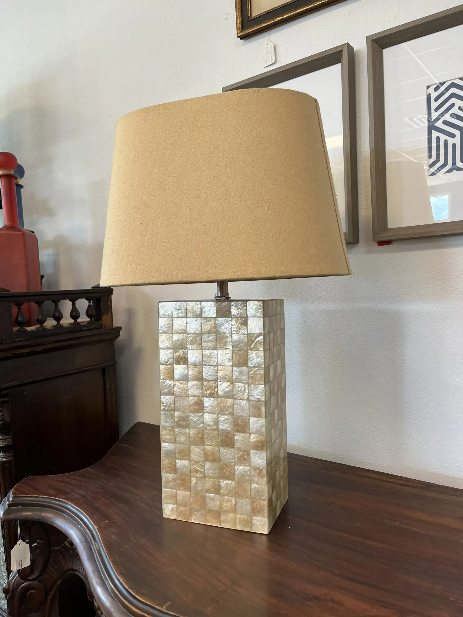 A rectangular lamp with a tiled mother-of-pearl base and a textured tan lampshade sitting on a dark wooden table.
