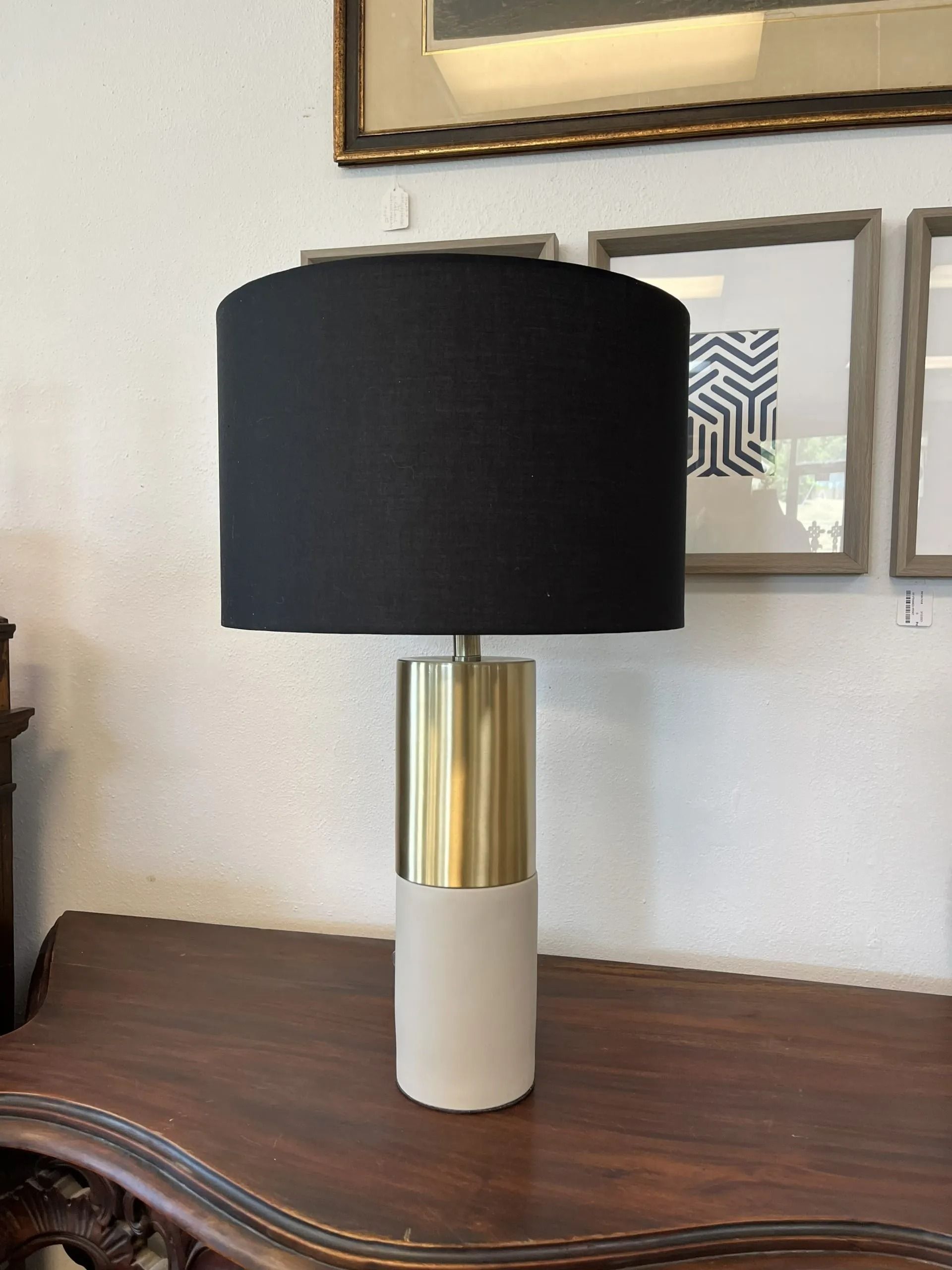 A table lamp with a black cylindrical shade, a gold metal top base, and a white concrete cylindrical bottom base.