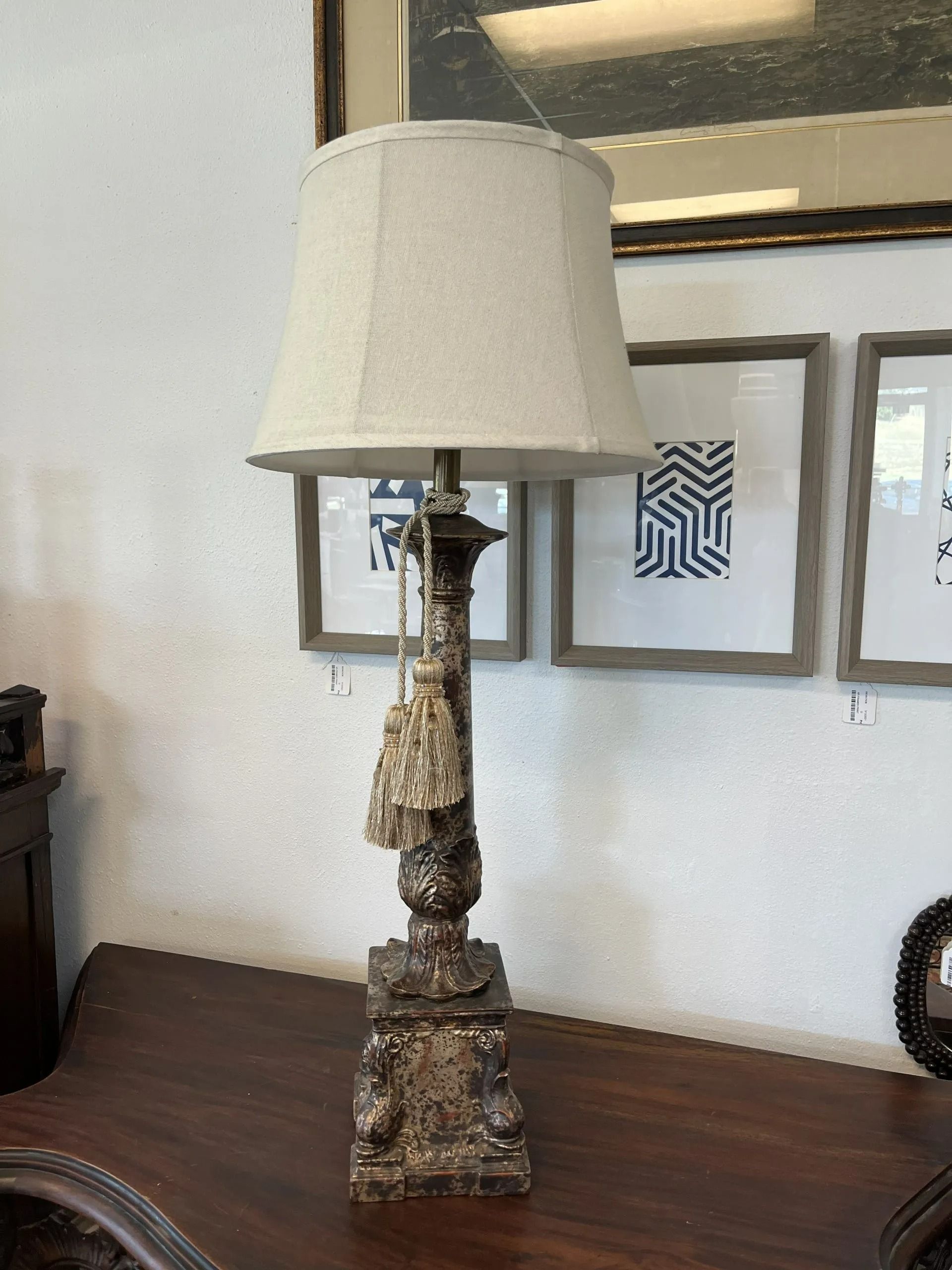 A rustic, brown ornate lamp with a beige fabric shade and a decorative tassel, sitting on a dark wooden table.