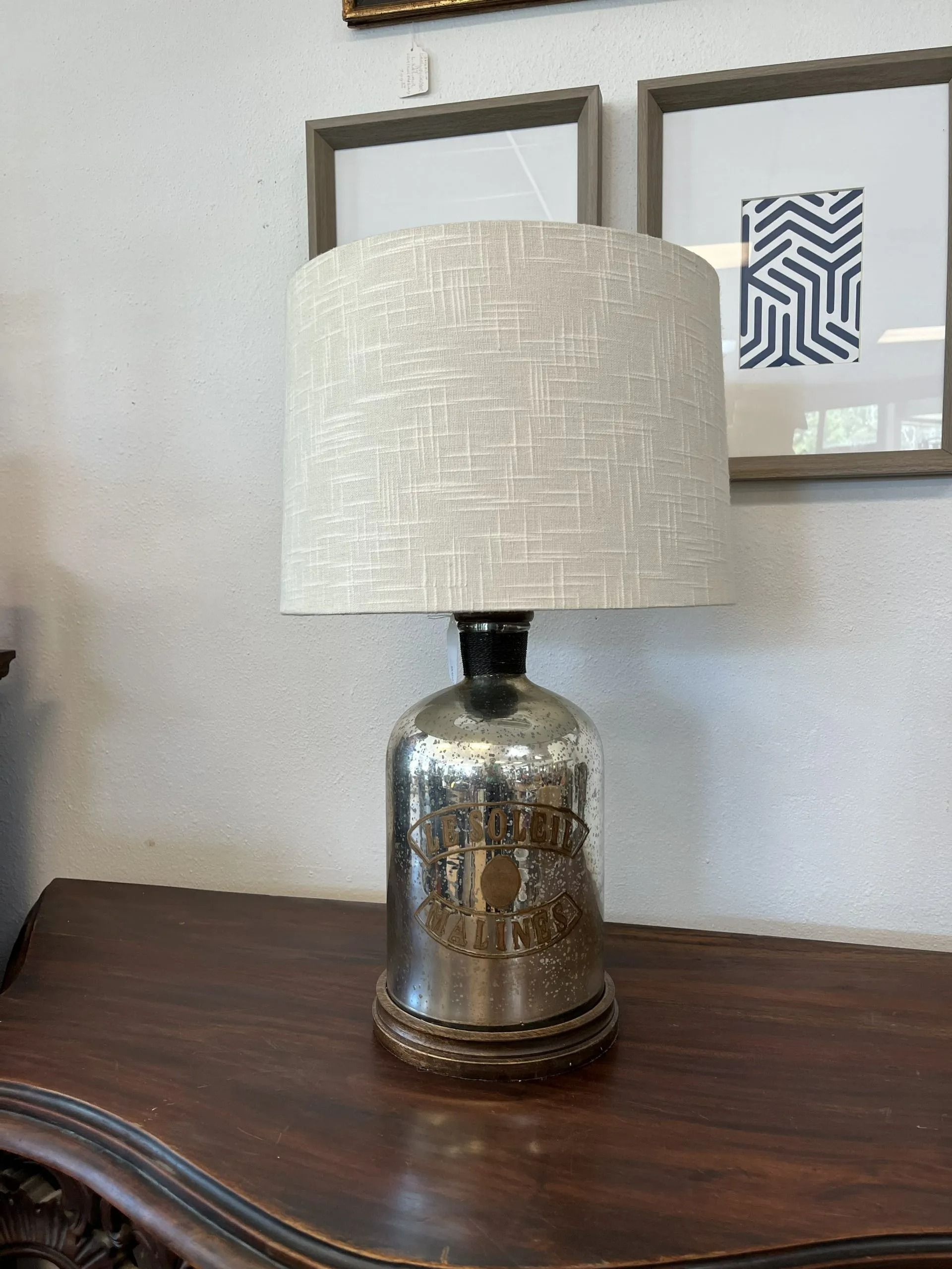 A table lamp with a mercury glass base and a light-colored, textured drum shade, sitting on a dark wooden surface.
