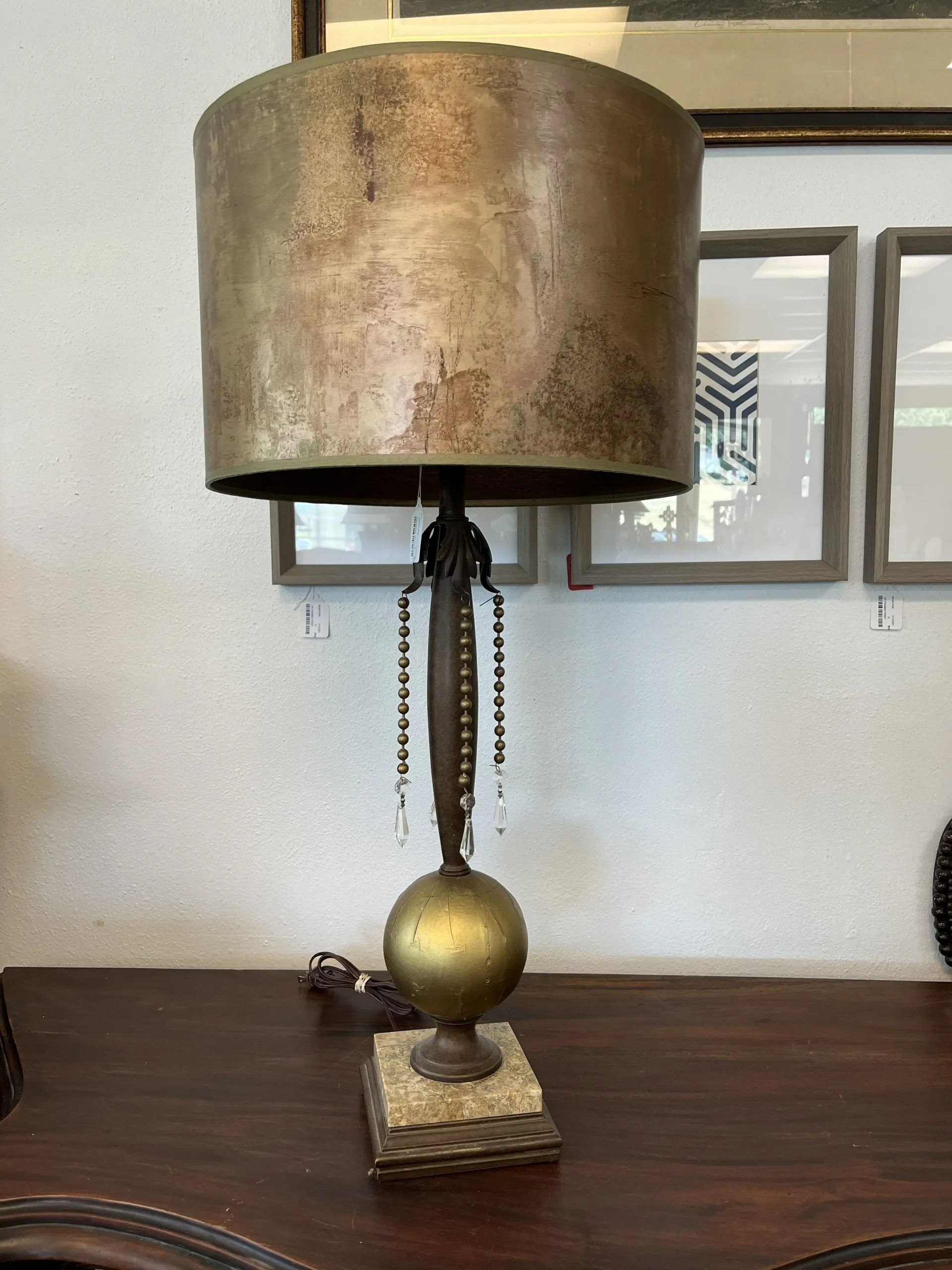A table lamp with a textured brass drum shade, a central post with hanging beads, a gold sphere, and a square base.