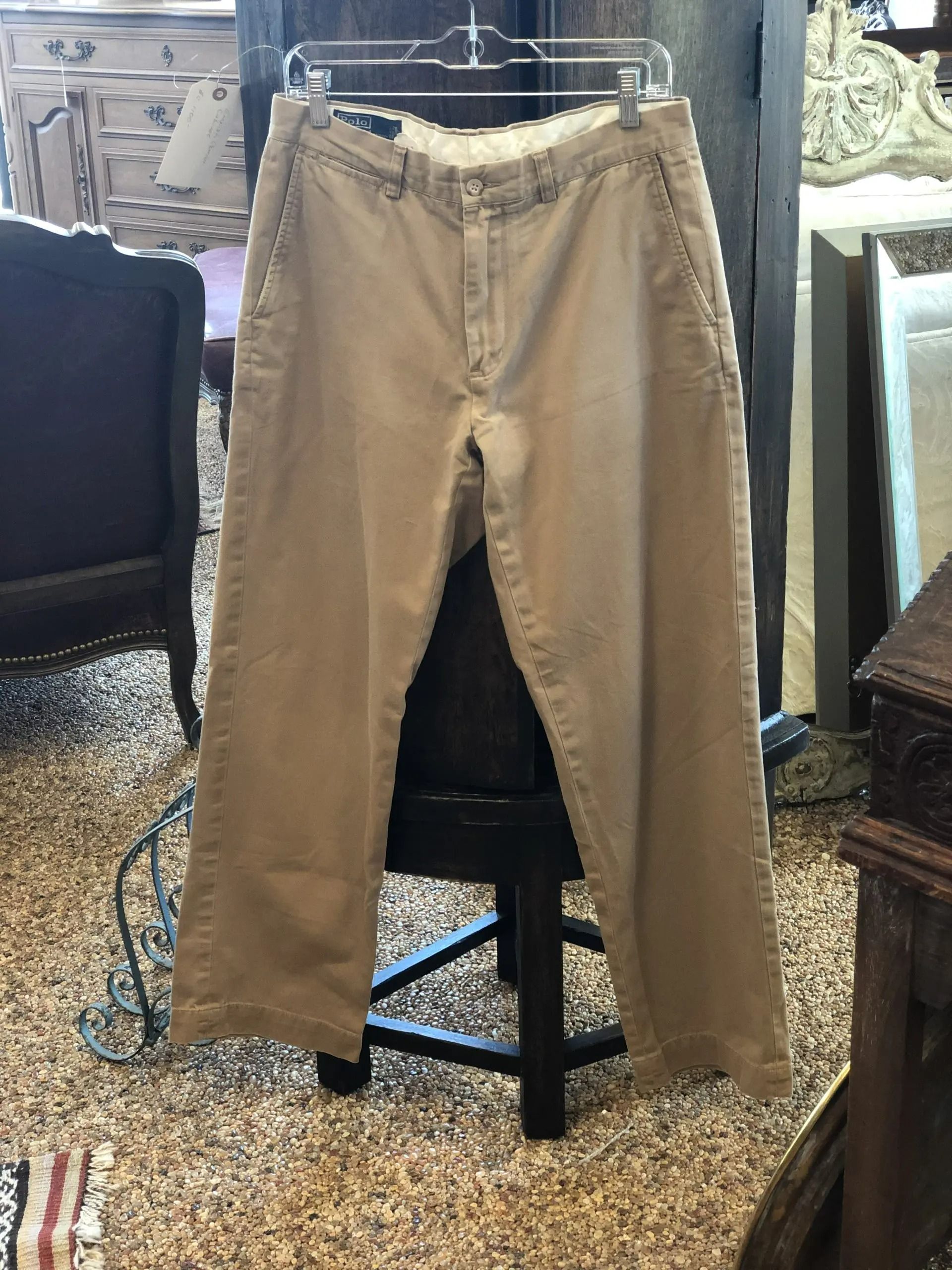 A pair of beige, straight-leg chino pants on a hanger, set against a background of furniture in a shop.