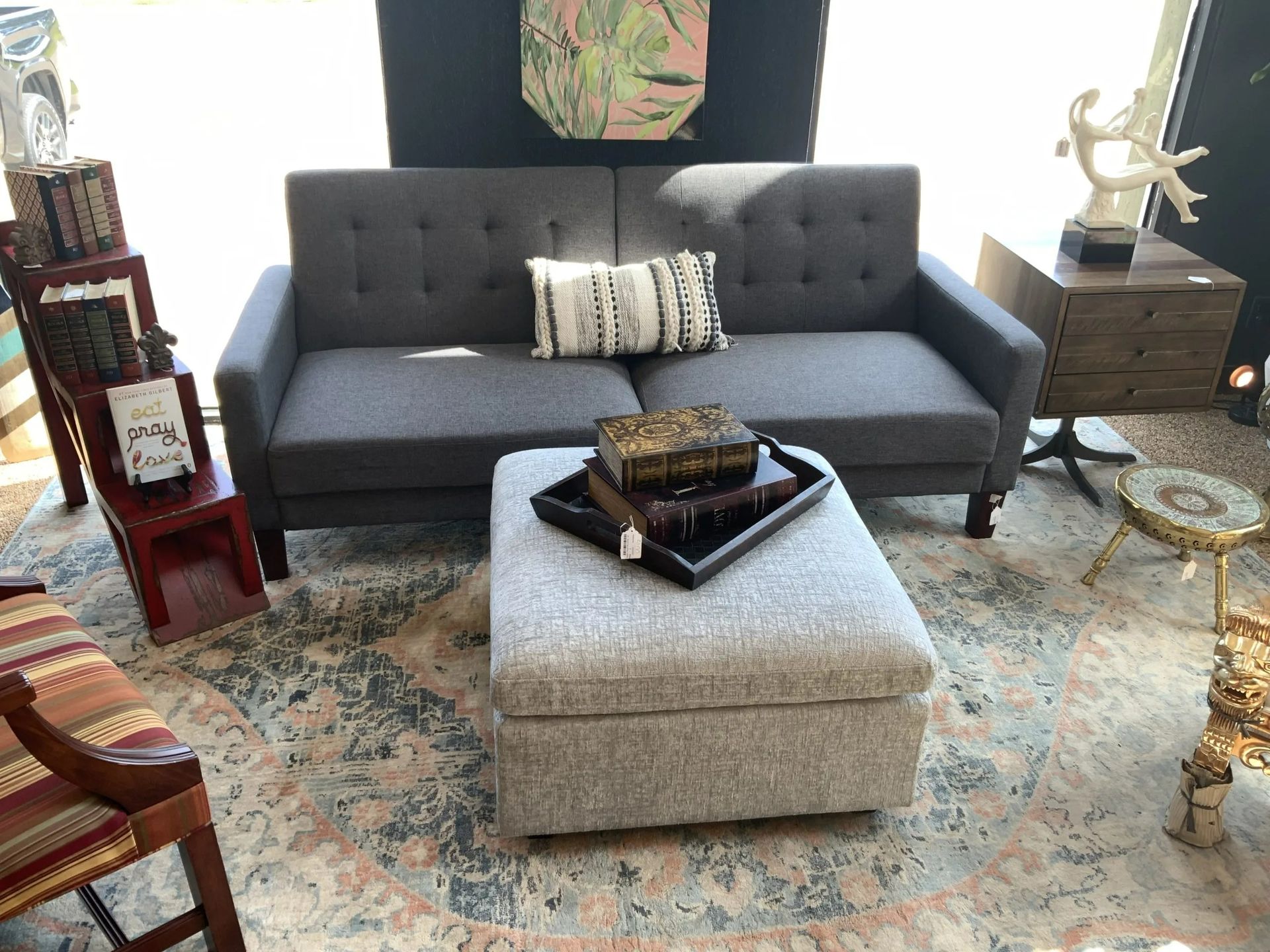 A gray tufted sofa with a patterned pillow, a square light gray ottoman, and decor arranged in a living room showroom.