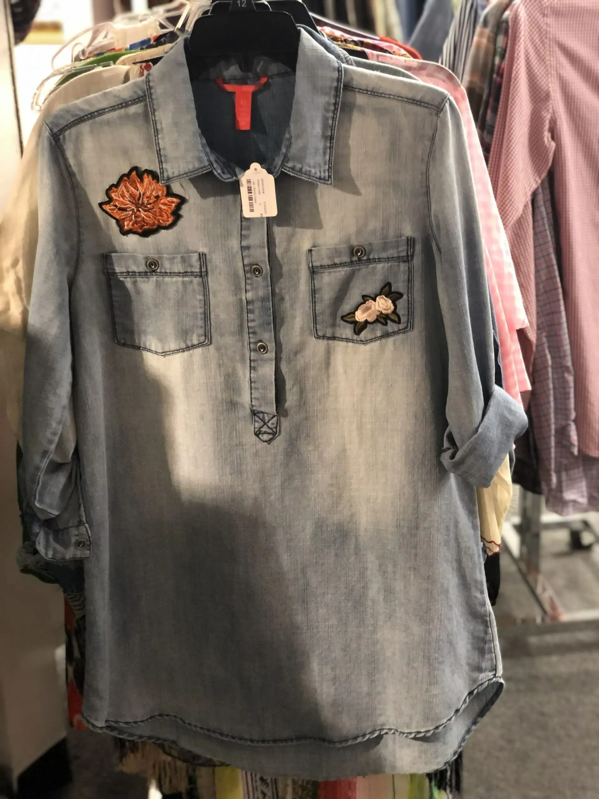 A long-sleeved, light-wash denim tunic with floral patches on the chest pockets, hanging on a clothing rack.