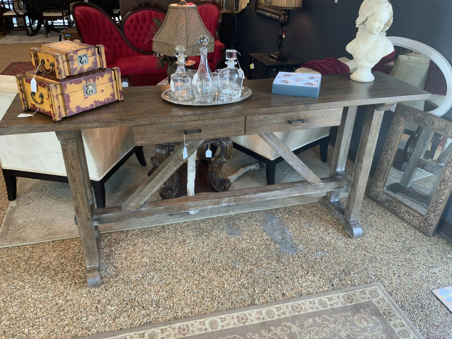 A rustic wooden console table with two drawers, topped with crystal decanters, decorative boxes, and a bust in a showroom.