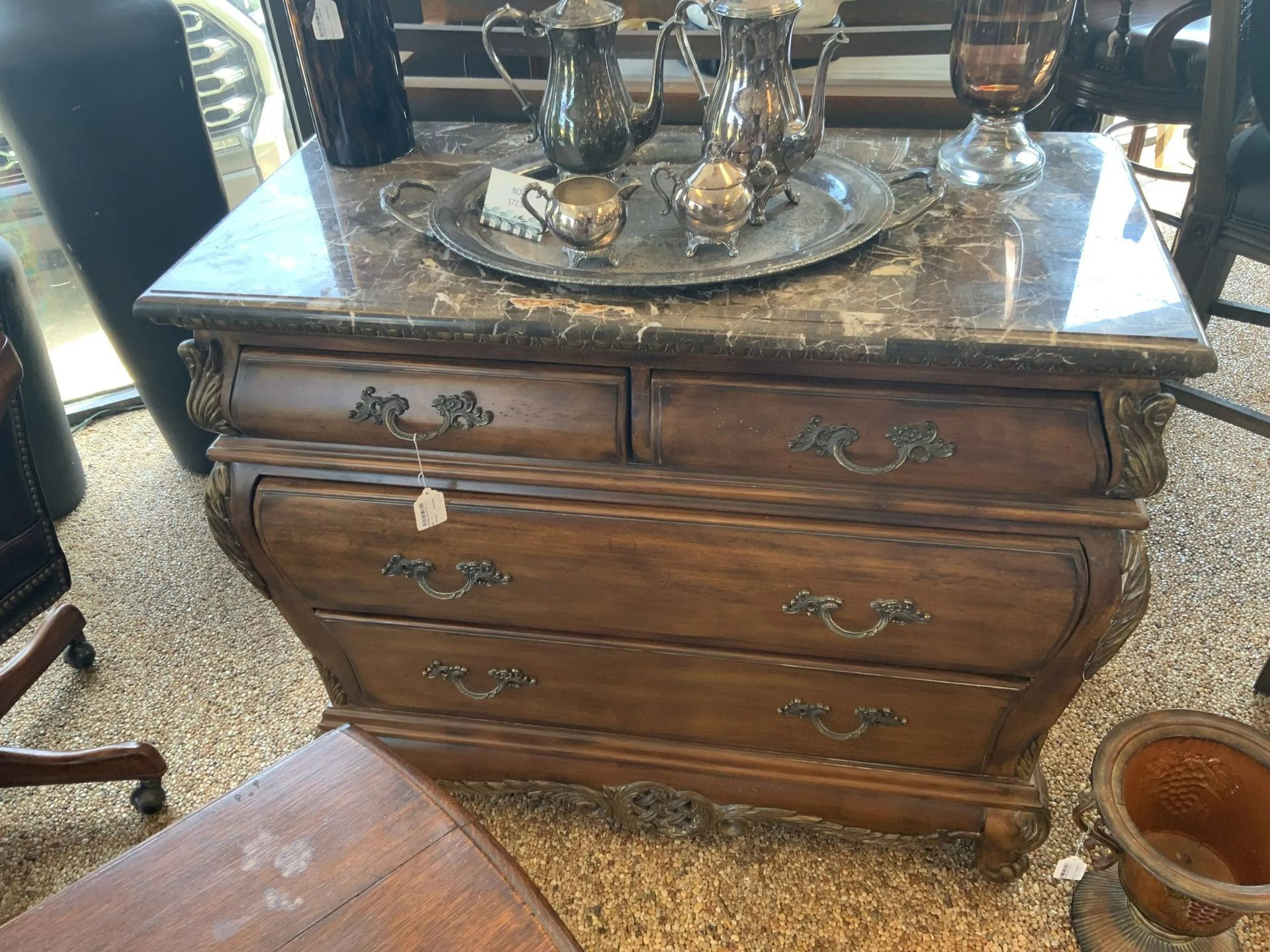 A wooden chest of drawers with a marble top, topped with a silver tea set, displayed in a store.
