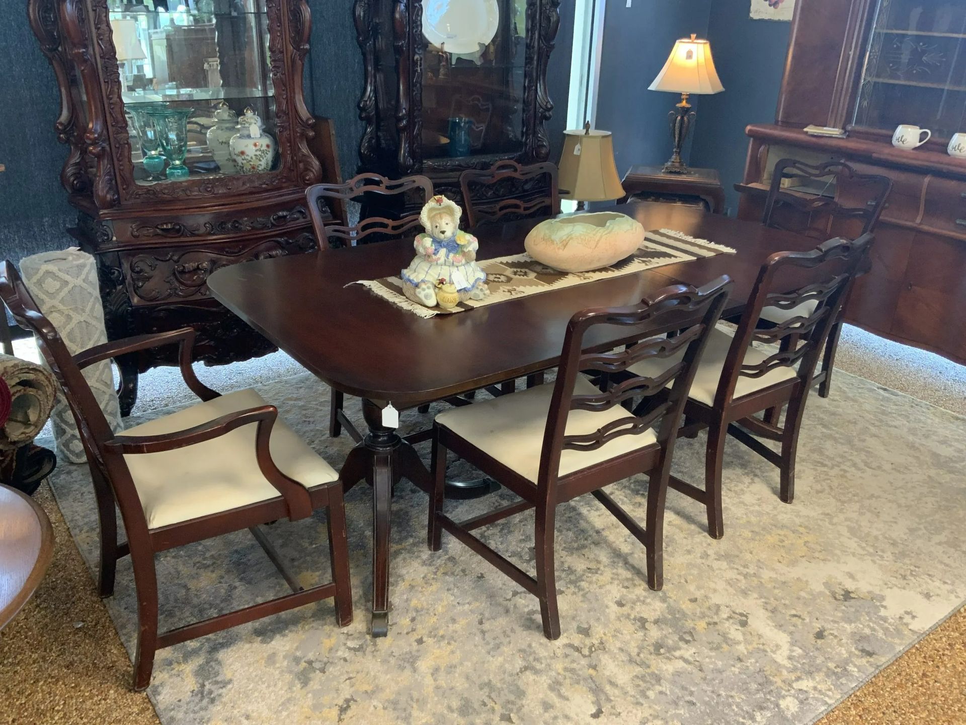 A dark wooden dining set with upholstered chairs sits on a patterned rug in a showroom filled with antique furniture.