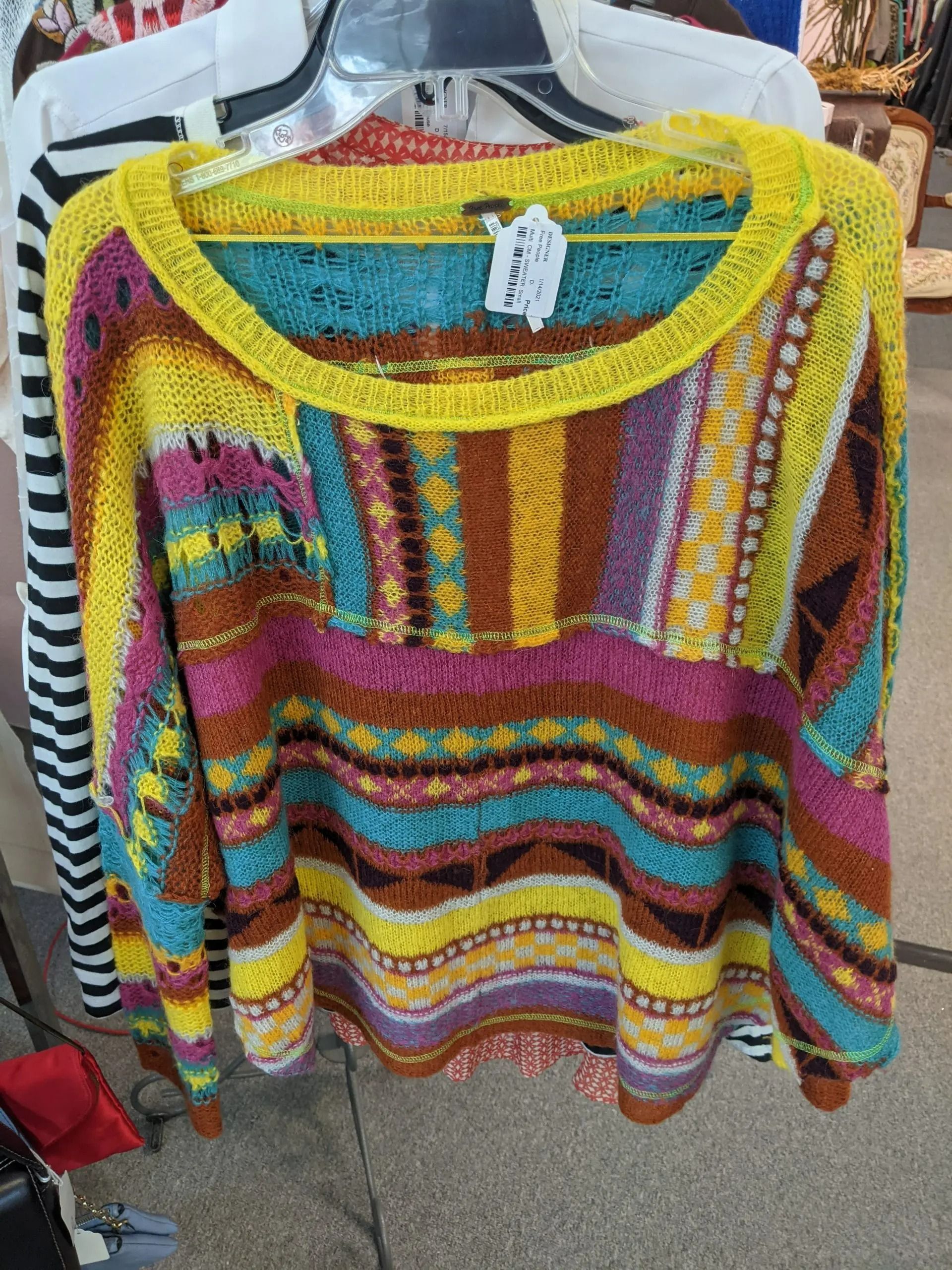 A colorful, loose-knit sweater with a horizontal patterned stripe design in yellow, teal, pink, and brown on a hanger.