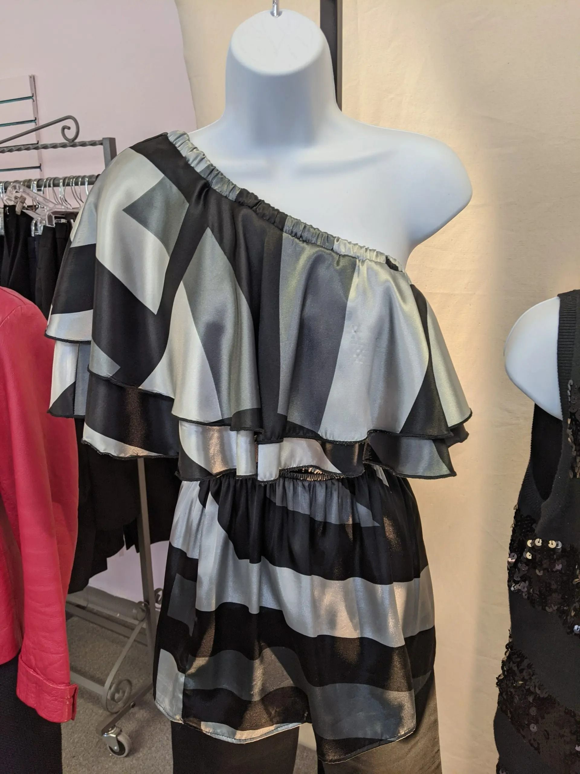 A mannequin wears a one-shoulder top featuring alternating black and silver horizontal stripes with ruffled sleeves.