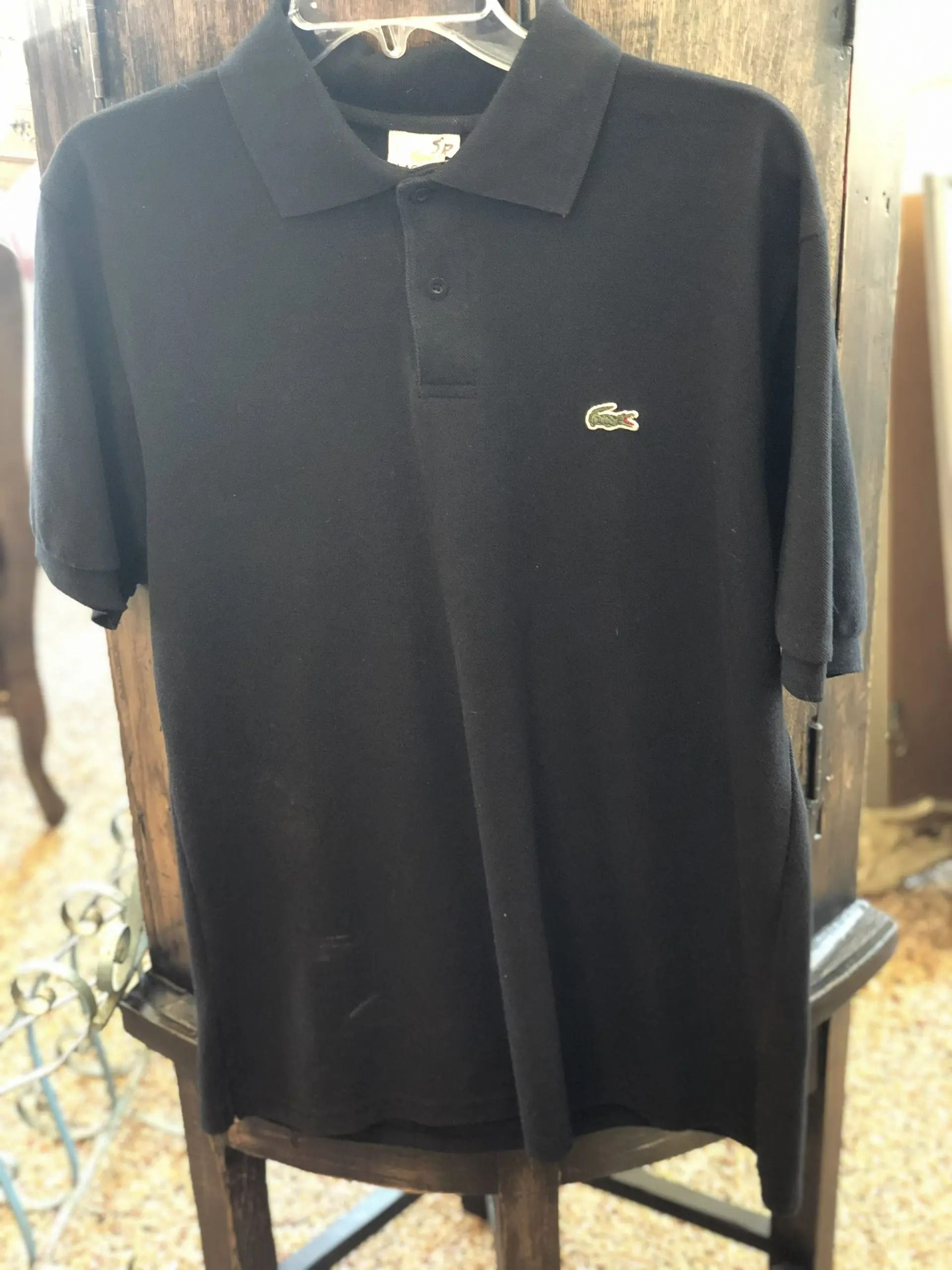 A black short-sleeved Lacoste polo shirt with a green crocodile logo, displayed on a hanger.