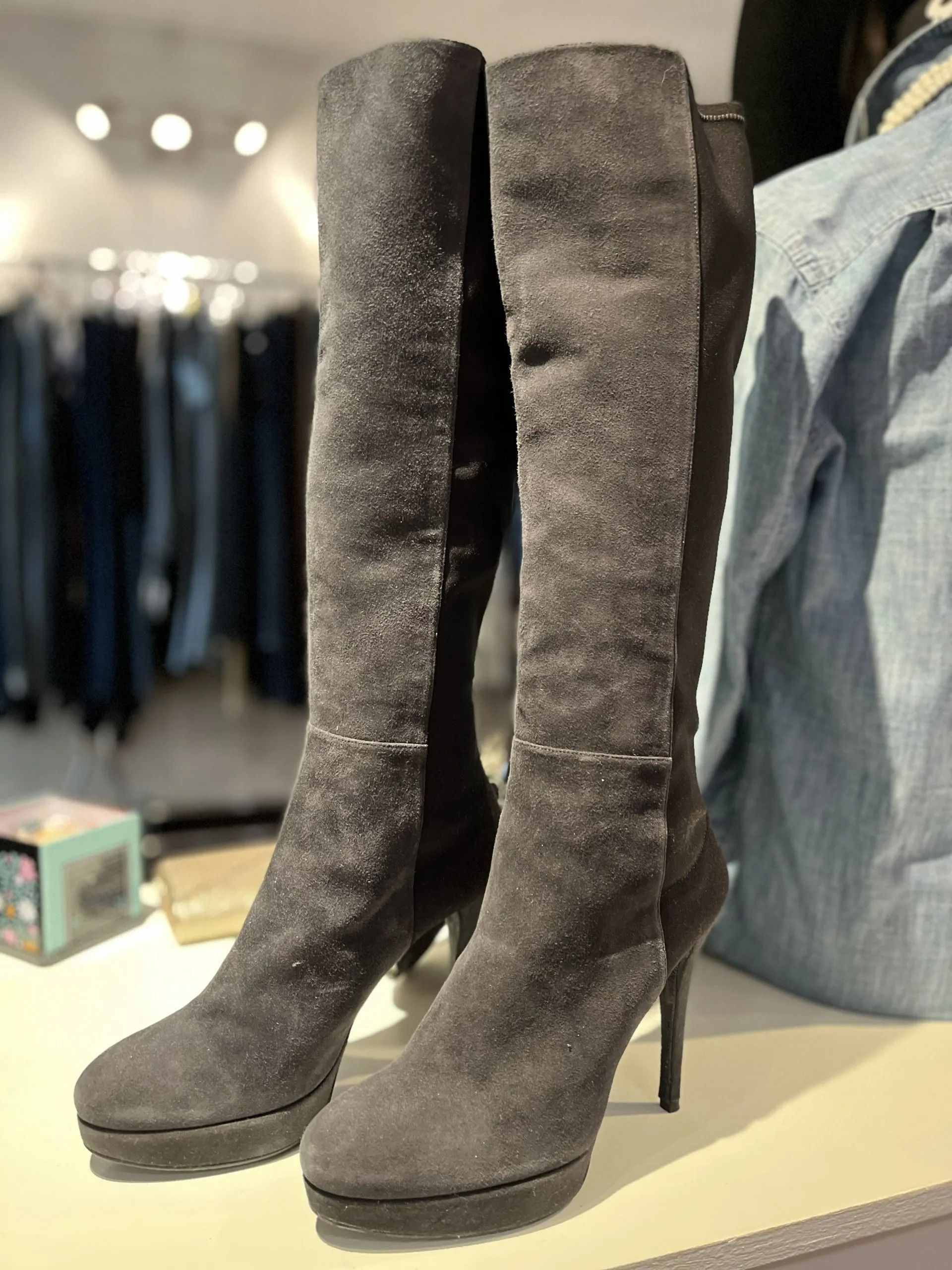 A pair of dark grey, knee-high suede boots with platform soles and thin heels displayed on a table in a retail store.