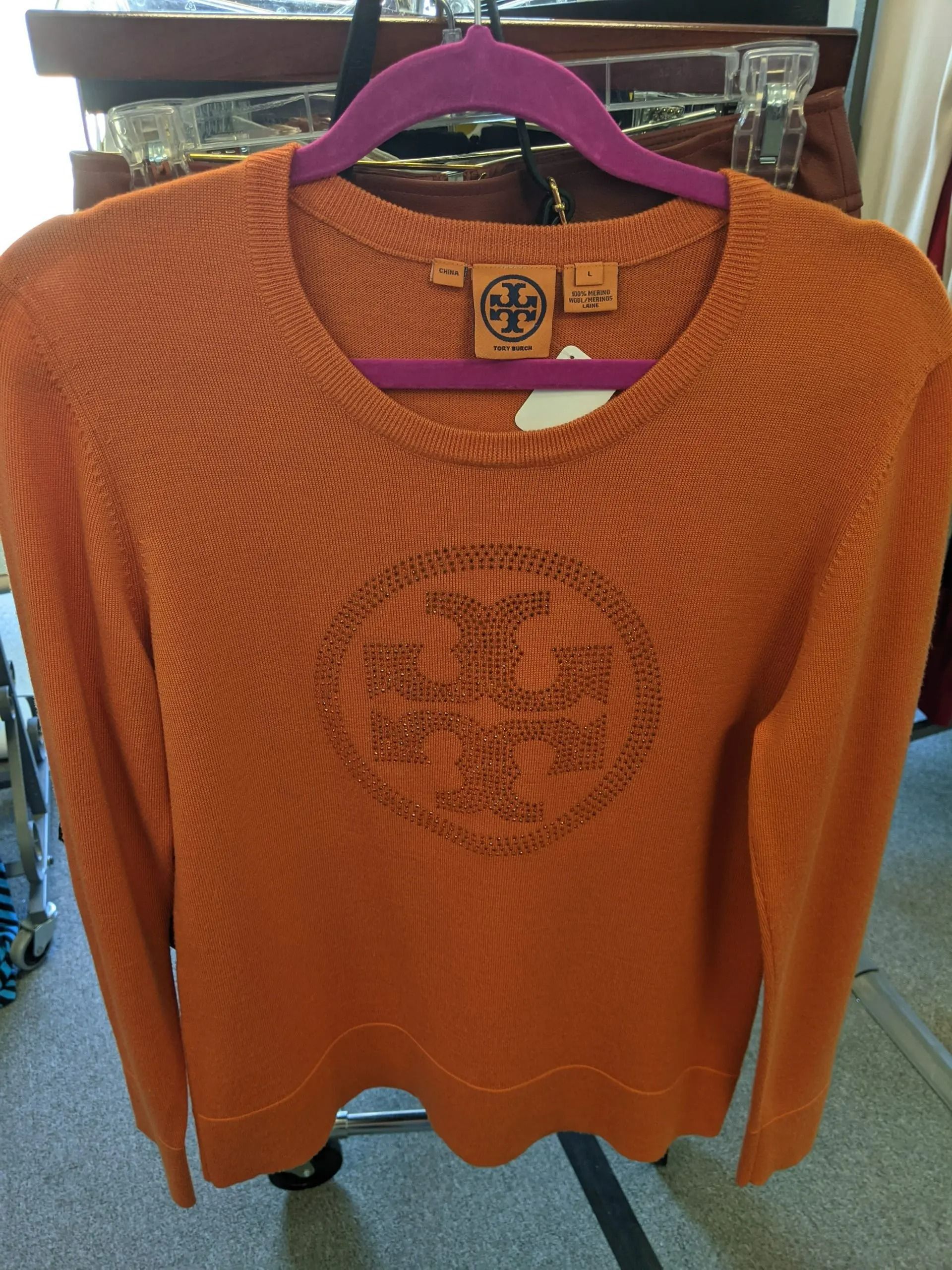 An orange long-sleeved Tory Burch knit sweater with a matching logo on the chest, hanging on a purple hanger.