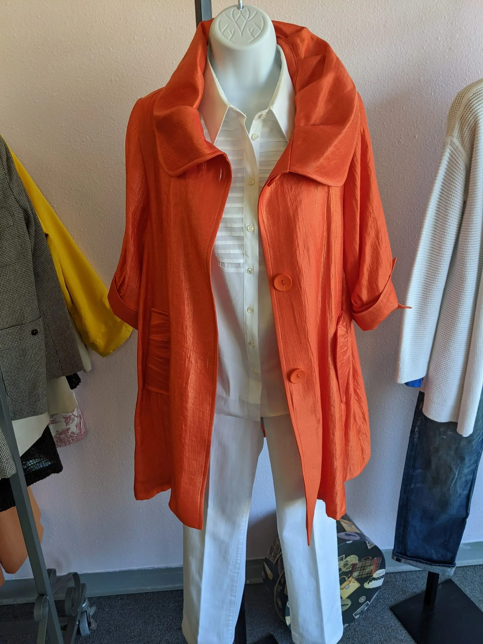 A bright orange, textured, buttoned jacket worn over a white collared shirt and white pants on a store mannequin.