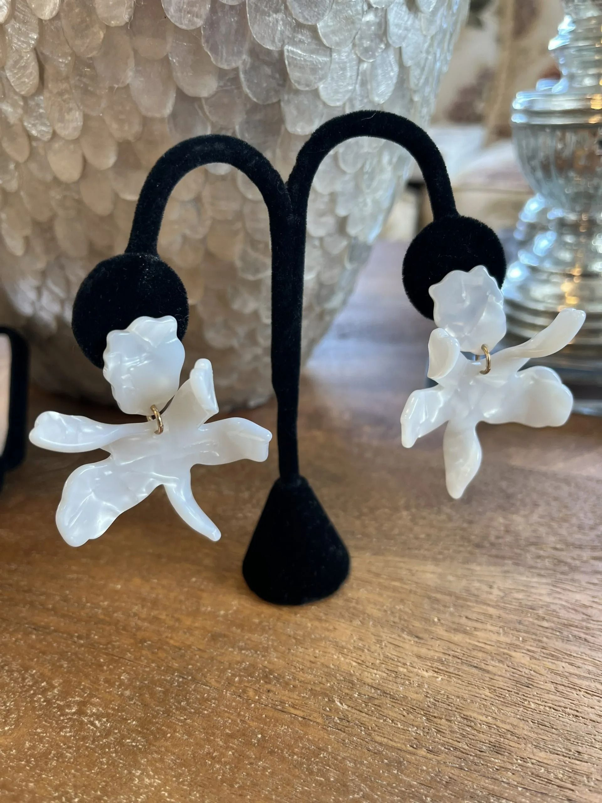 A pair of white, translucent, orchid-shaped earrings displayed on a black stand atop a wooden surface.