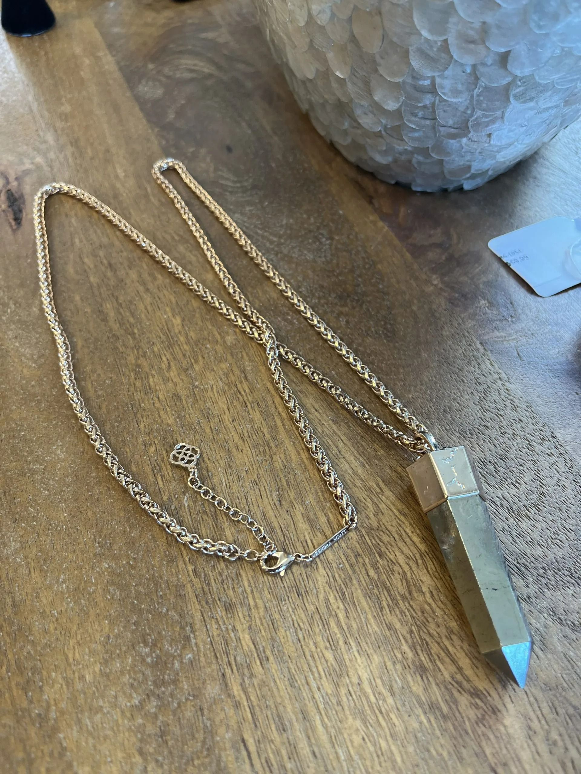 A gold-toned, faceted crystal pendant on a delicate chain, resting on a wooden surface next to a white textured object.