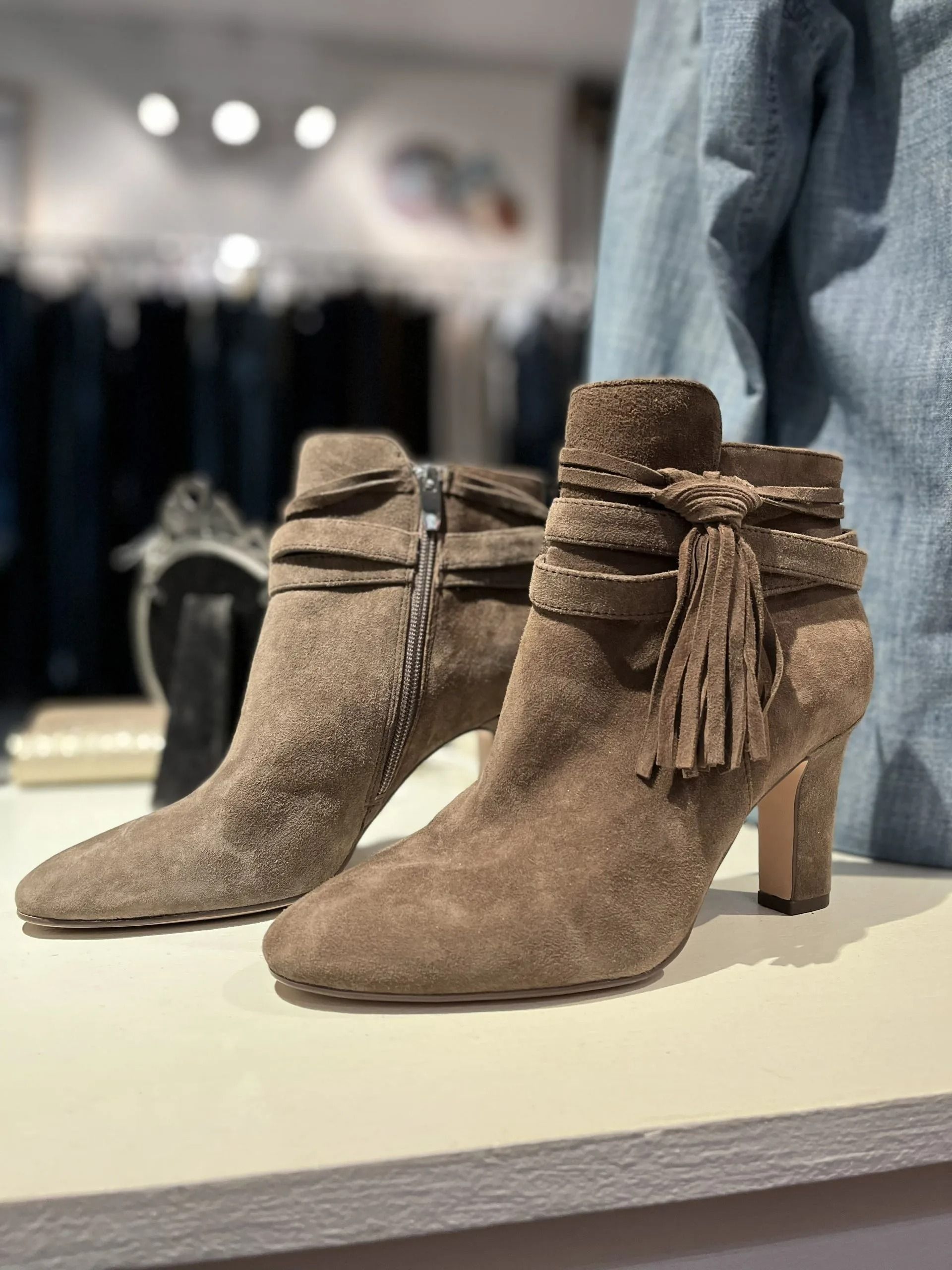 Two taupe suede ankle boots with fringe tassels and wrap-around straps, displayed on a store shelf.
