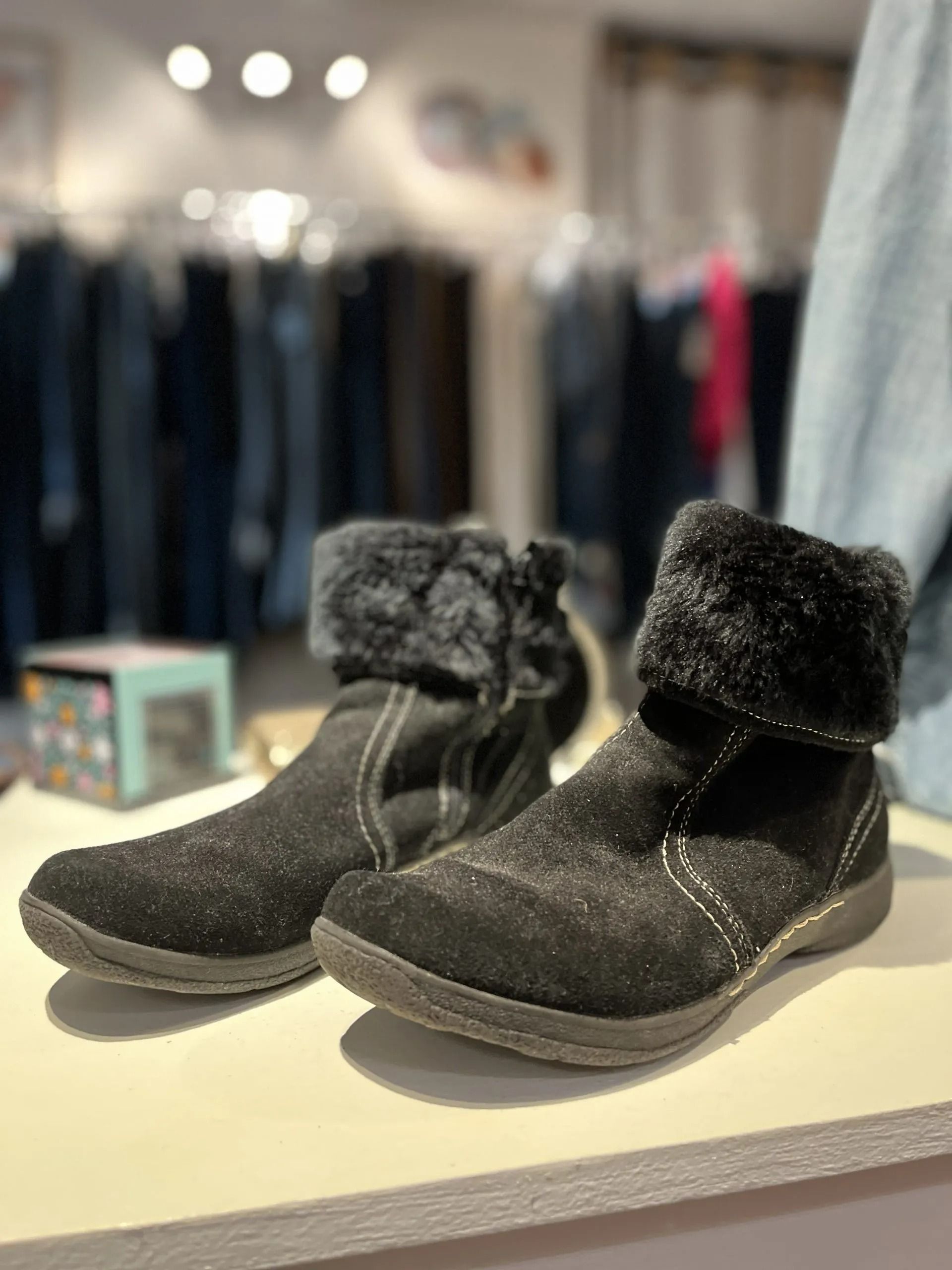 A pair of black suede ankle boots with faux fur cuffs displayed on a white shelf in a clothing store.