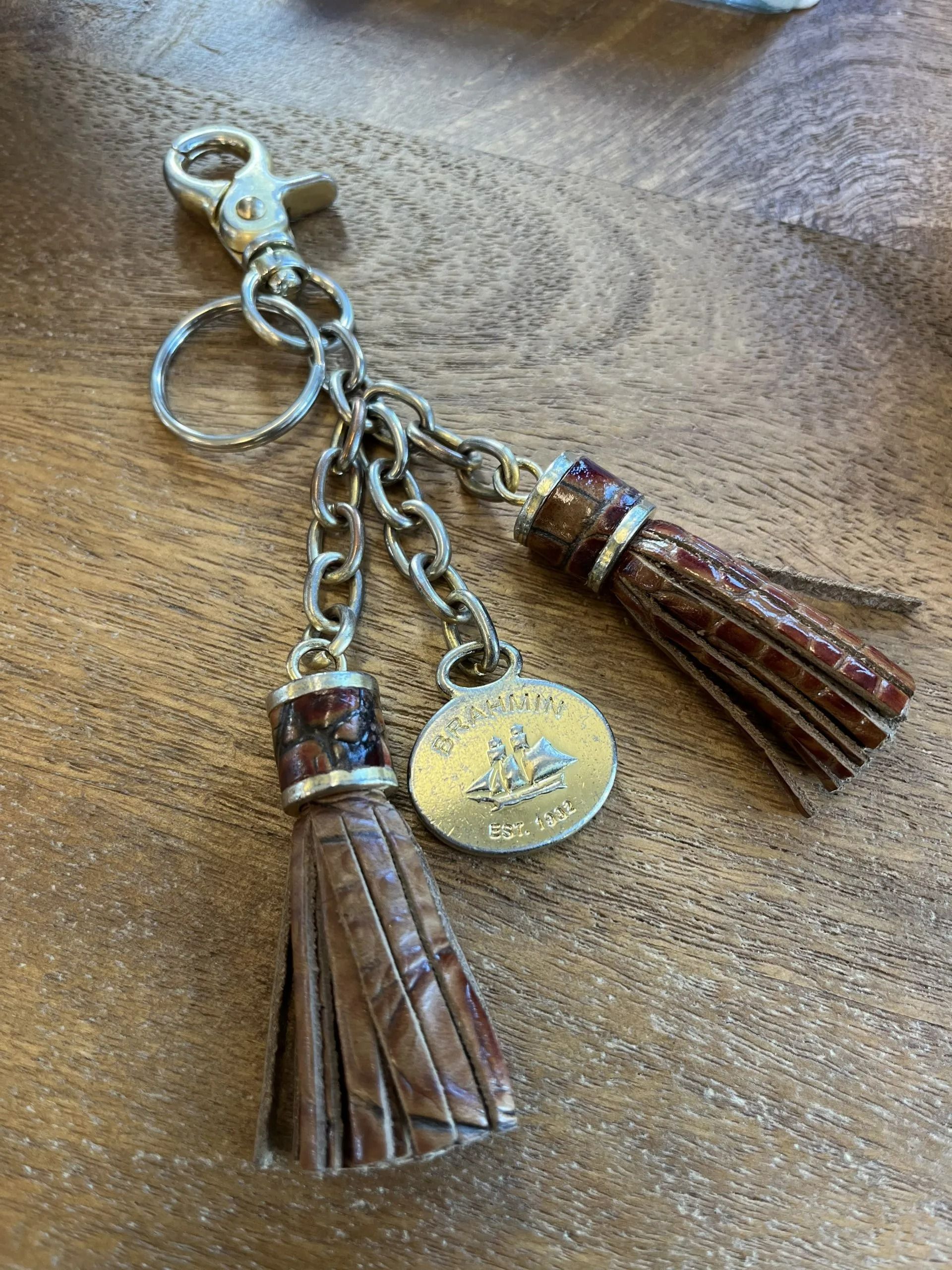 A brown leather tassel keychain with a circular metal charm, hanging from a metal clip against a wooden background.