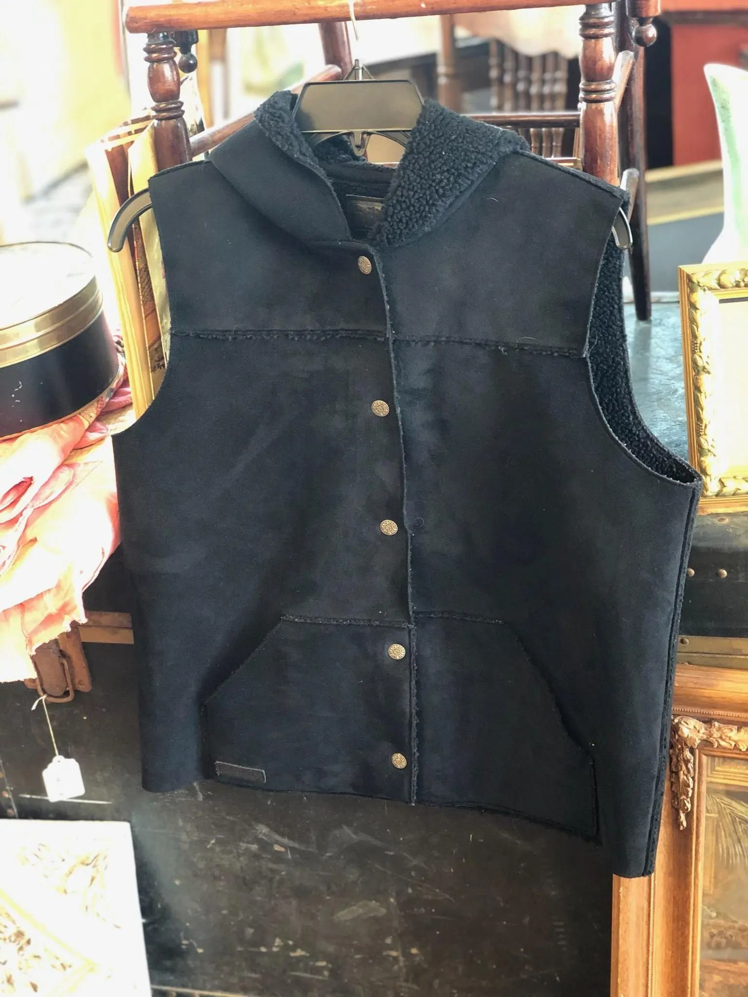 A black suede, hooded vest with button closures, hanging on a hanger against a blurred background.