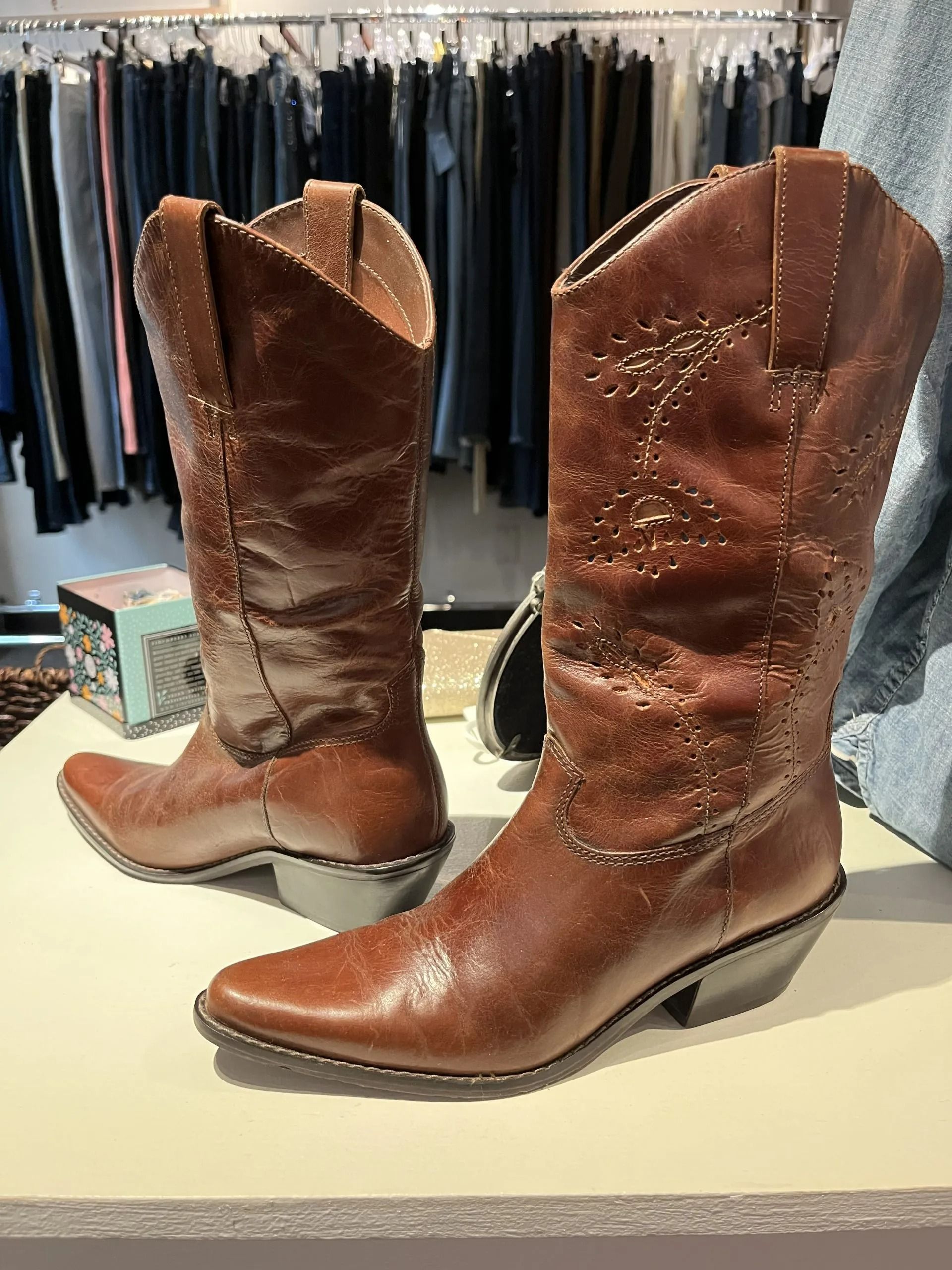 A pair of brown, pointed-toe cowboy boots with decorative stitching, displayed on a surface in a clothing store.