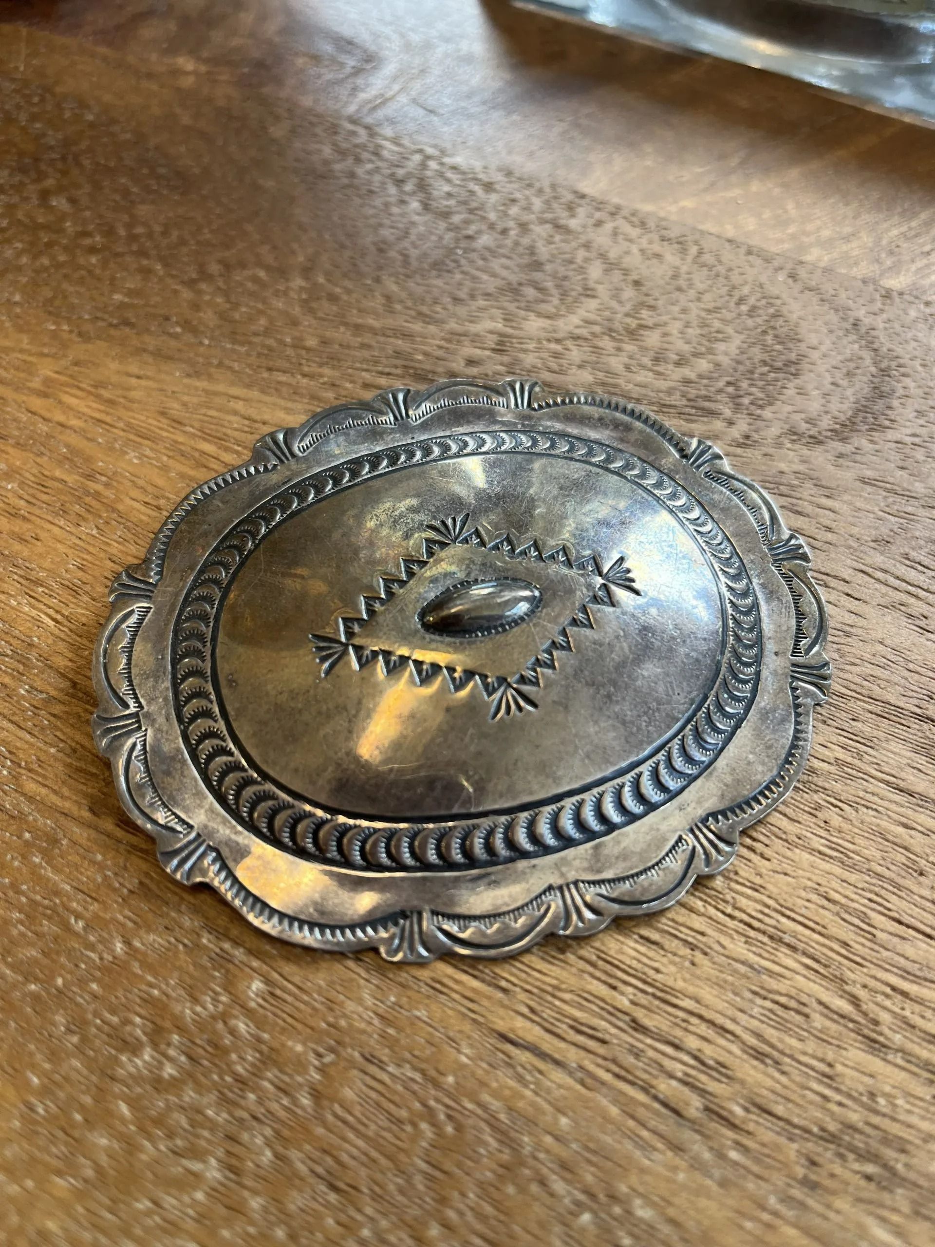An oval, tarnished silver decorative buckle featuring a central diamond pattern, resting on a wooden surface.
