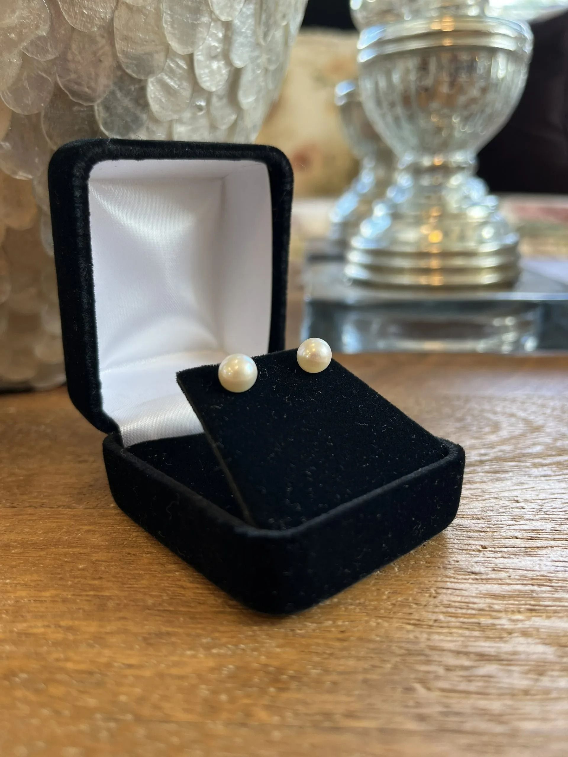 A pair of white pearl stud earrings sits in an open black jewelry box on a wooden surface.