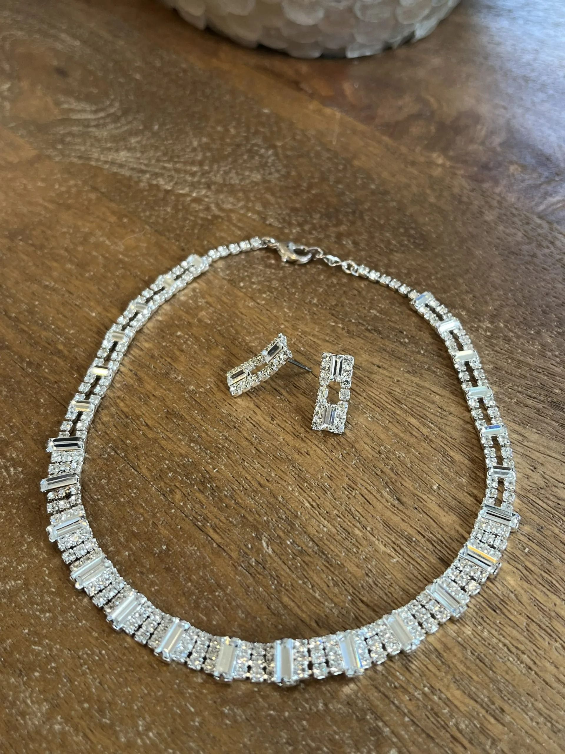 A silver-toned necklace and matching pair of stud earrings, decorated with baguette and round crystals on a wood surface.