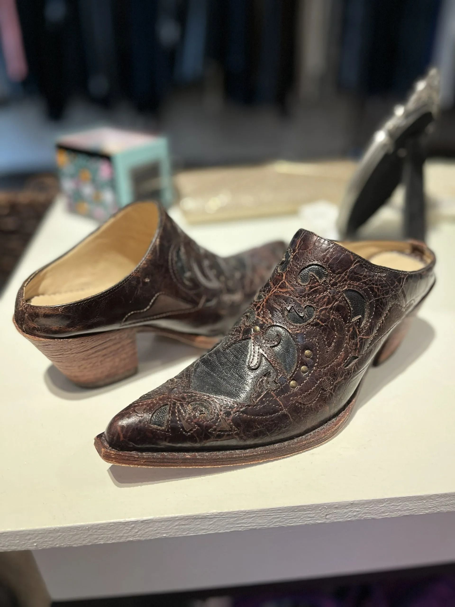 Two dark brown leather mule-style shoes with western-inspired embossed detailing and wooden heels on a white surface.