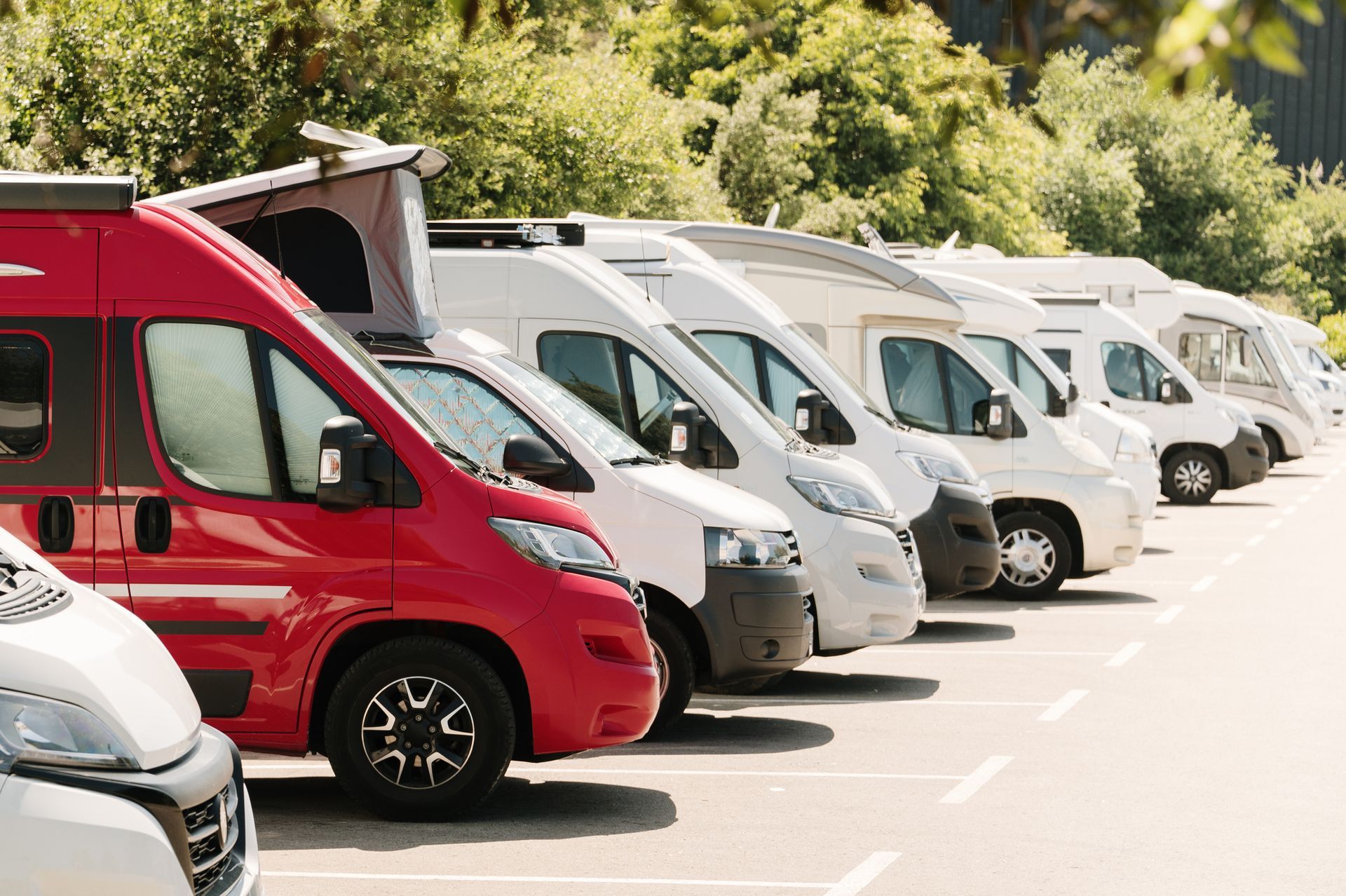 A row of camper vans parked outside; red, white and silver vehicles face the same direction in a parking lot.