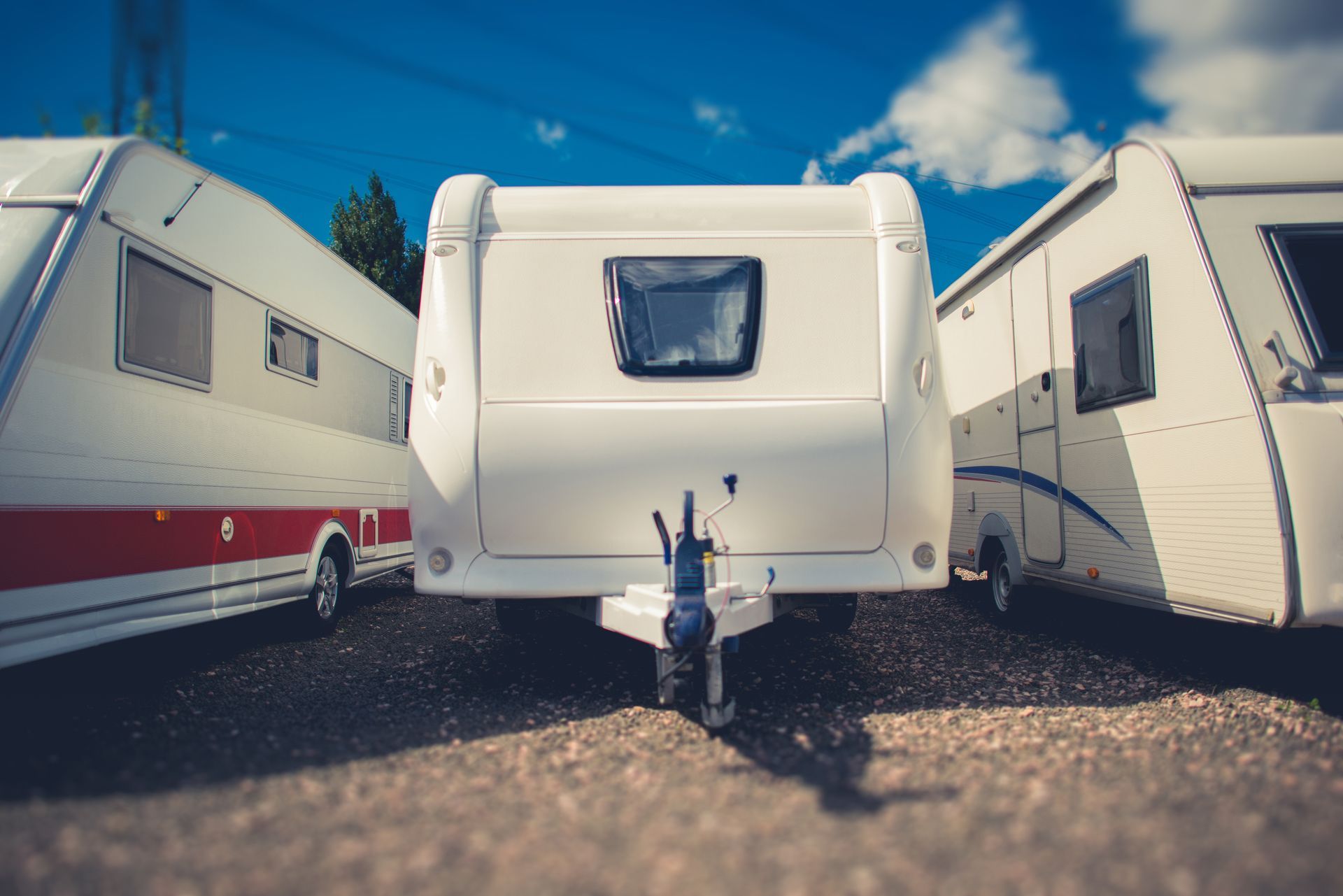 Three travel trailers parked on a gravel lot under a blue sky with clouds.