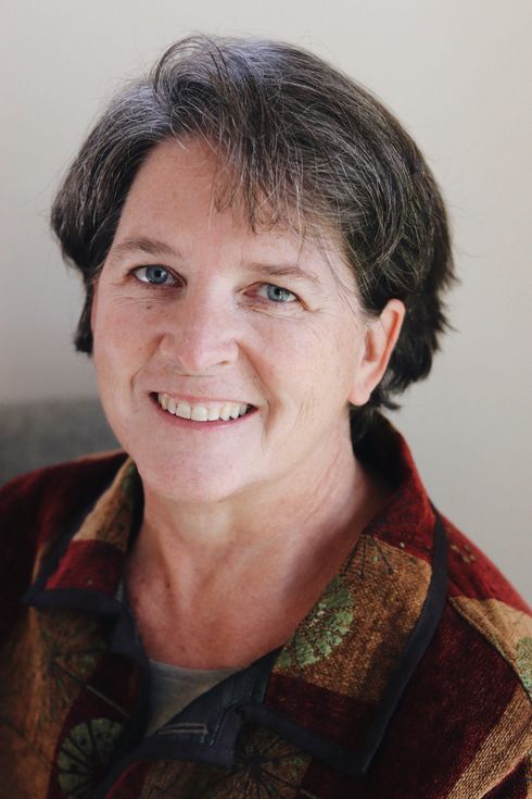 Woman smiling, wearing a colorful, patterned button-down shirt. Short, dark hair and blue eyes.