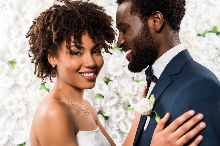 Smiling couple; bride in white dress, groom in suit, in front of a white floral wall.