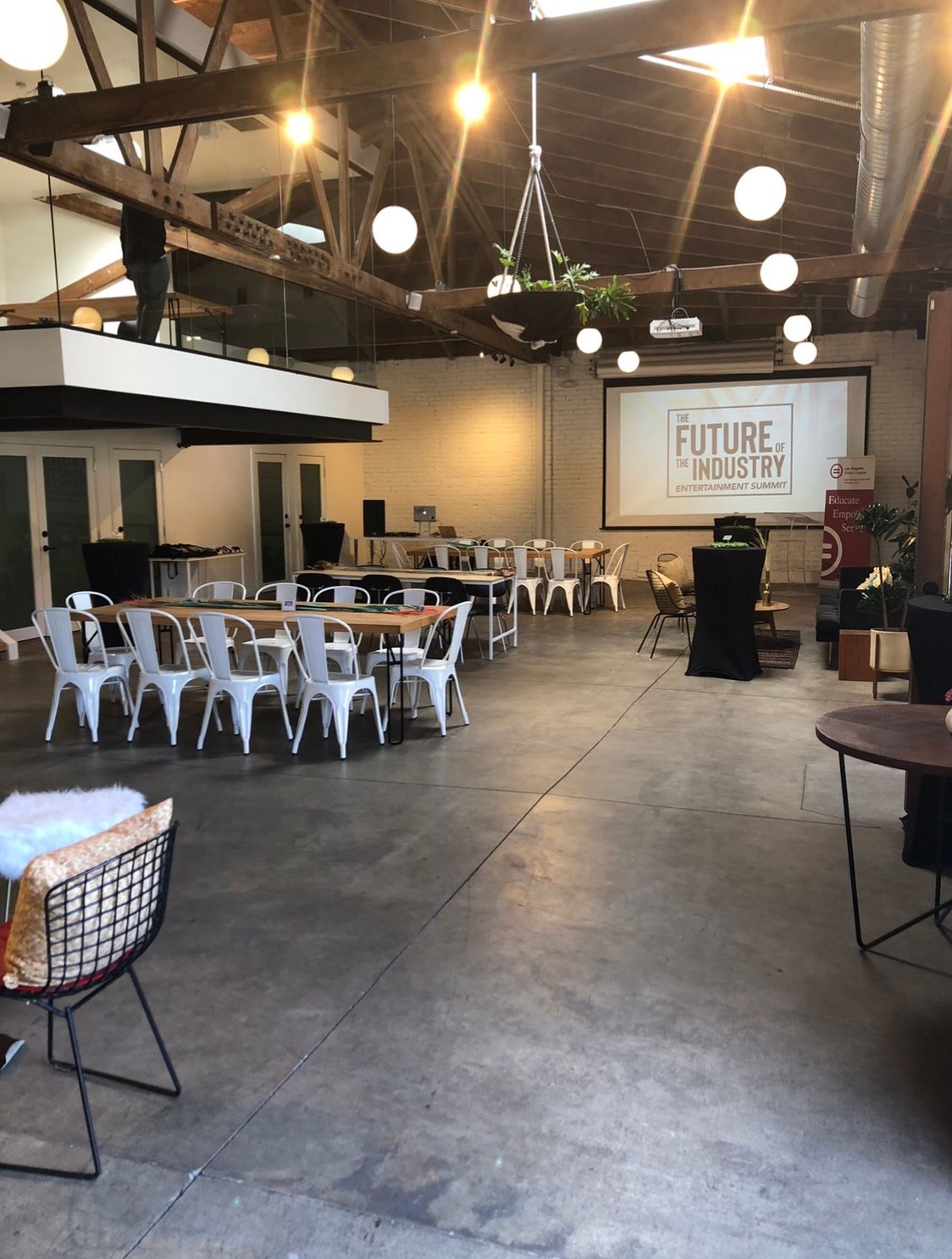 Spacious event hall, concrete floor, tables, chairs, projector screen, hanging plants, overhead lights.