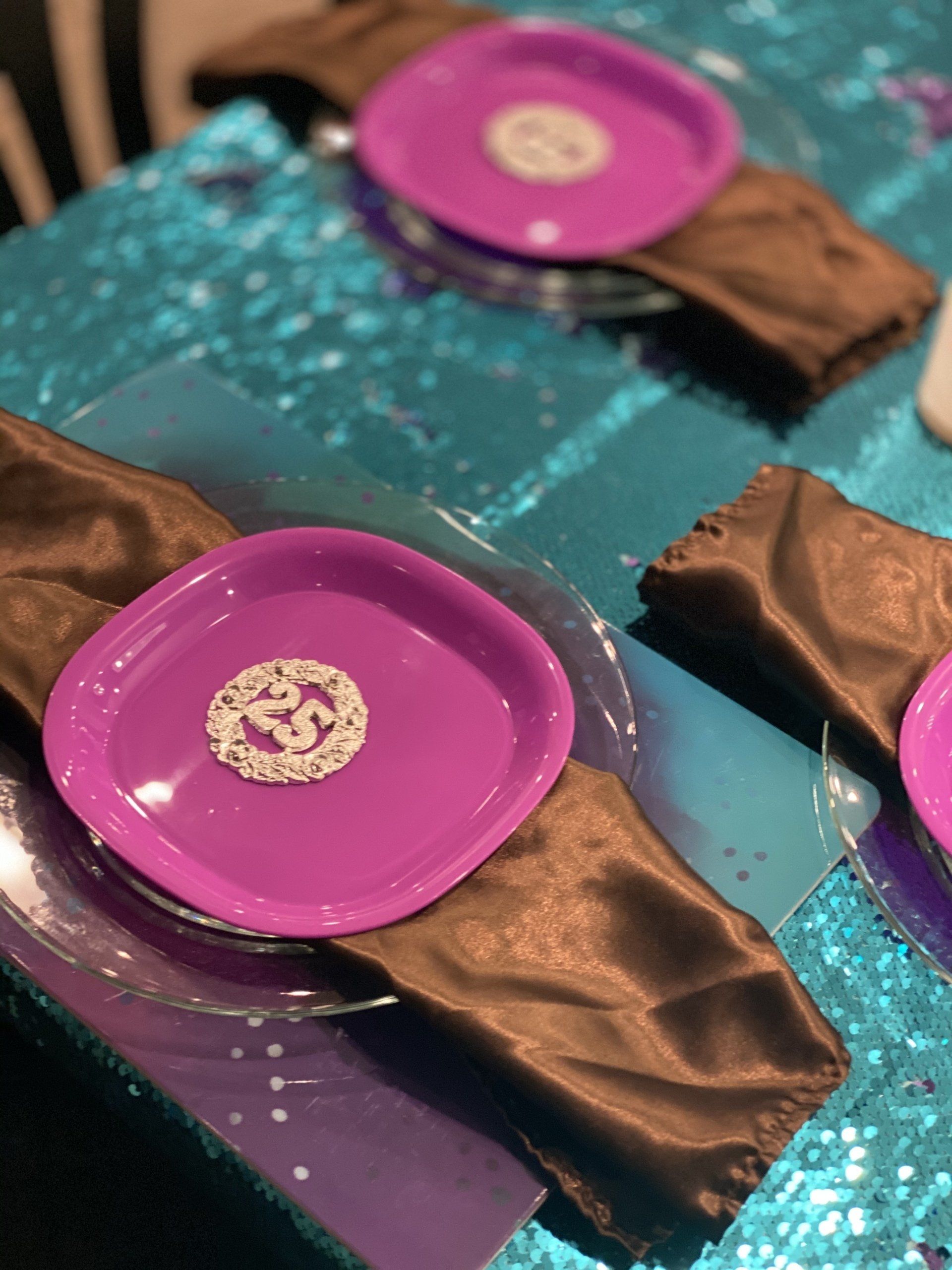 Purple plates with white detail atop brown cloth napkins on a teal tablecloth.