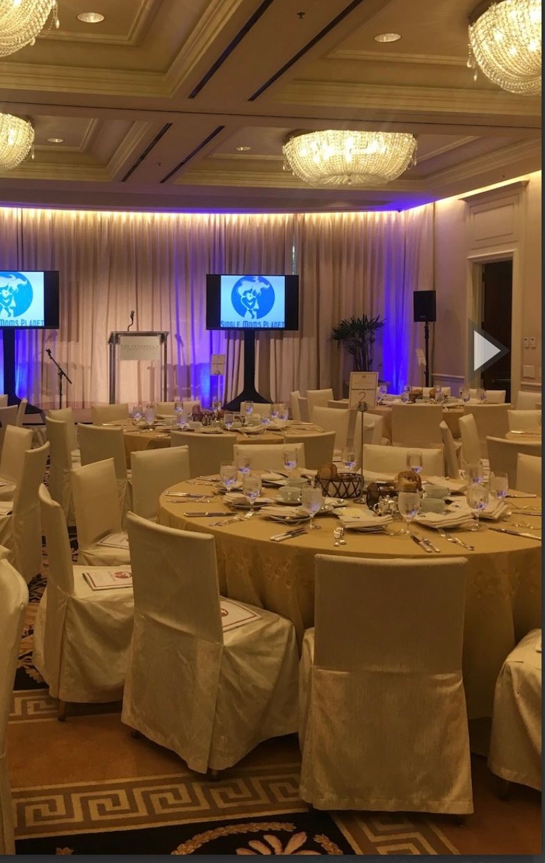 Formal banquet hall set for an event; round tables with white chairs and gold tablecloths. Two screens and podium.