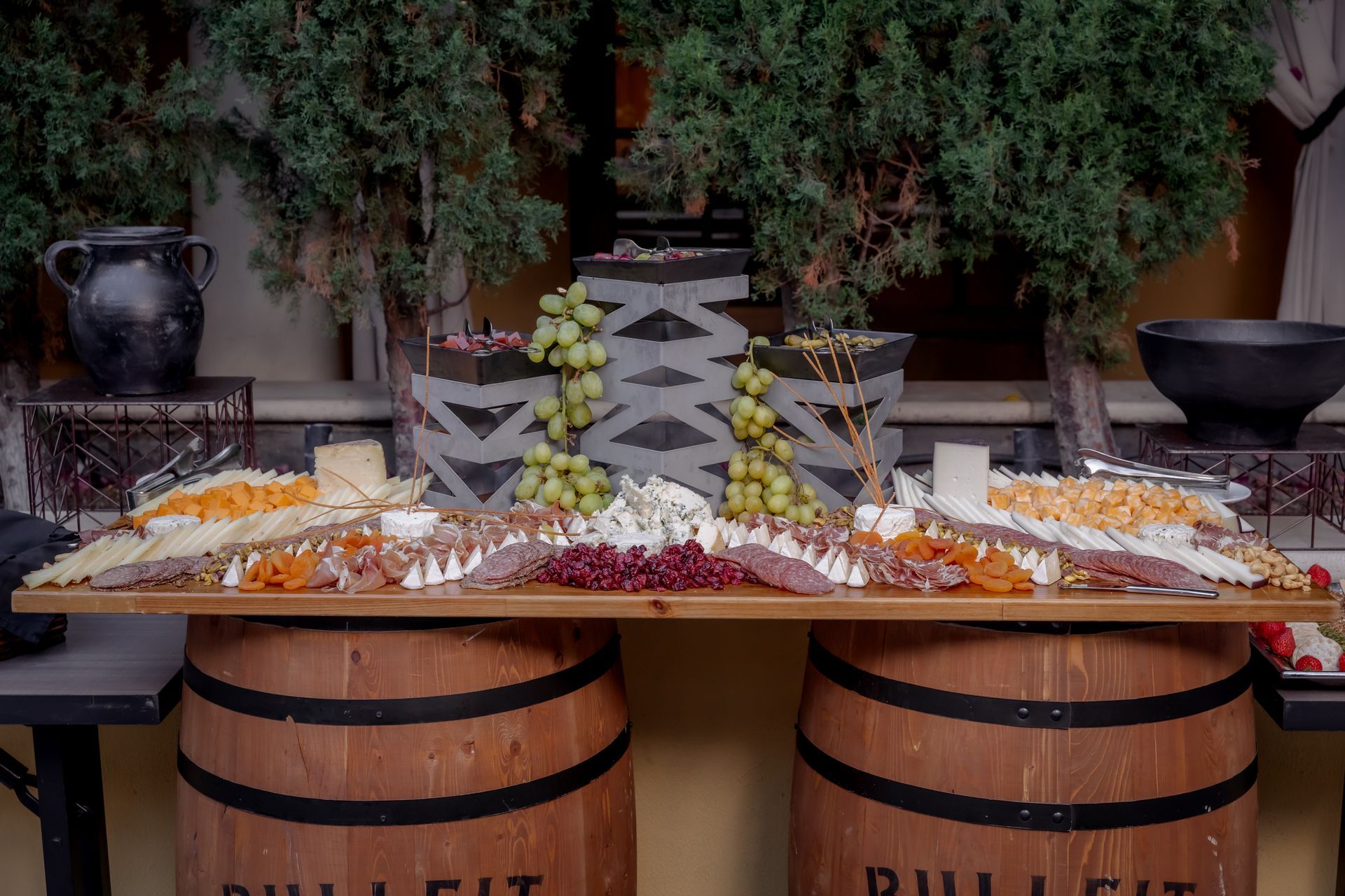 Charcuterie board on barrels with grapes, cheese, and meats, set in front of greenery.
