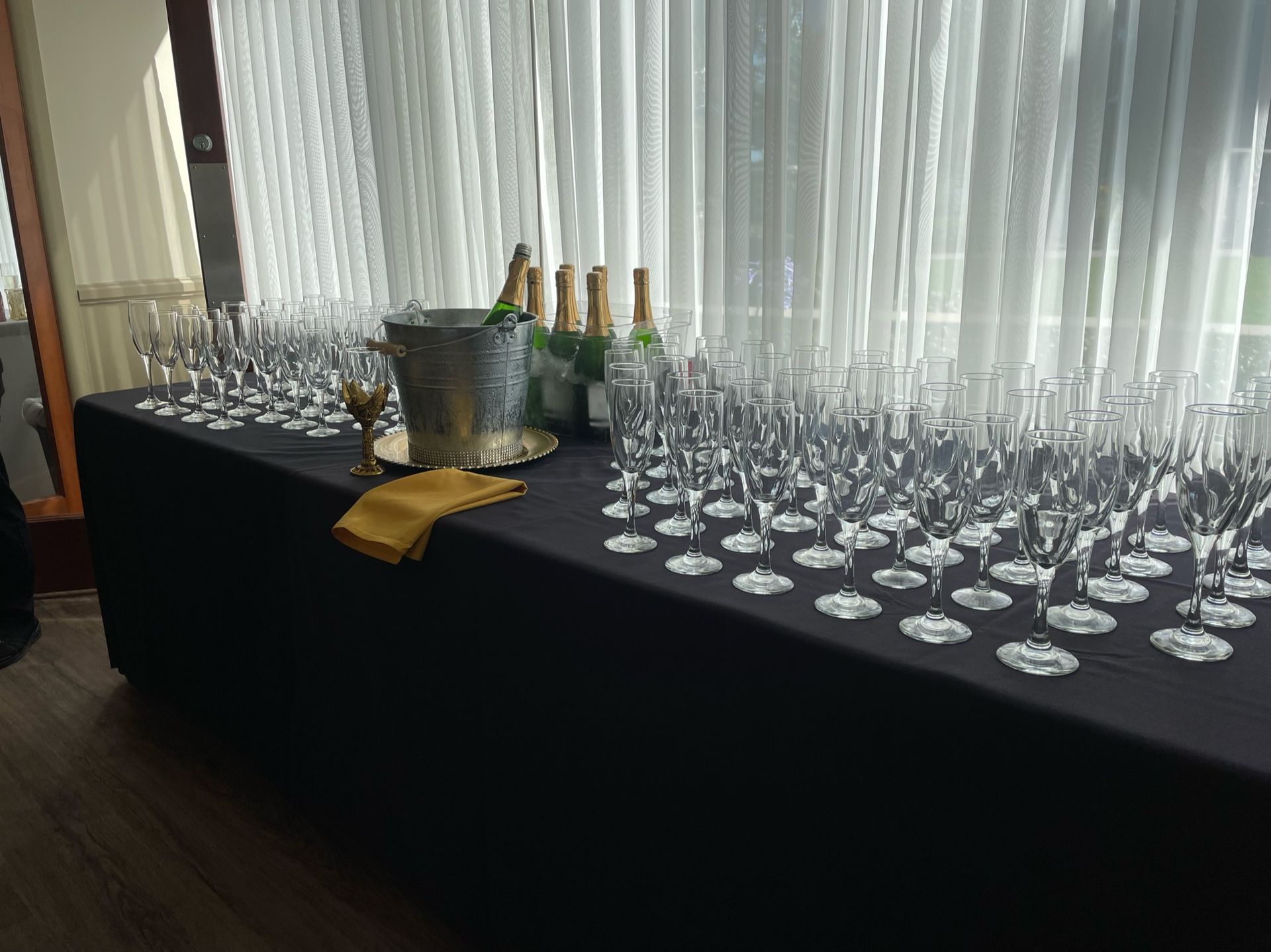 Table set for a celebration with champagne bottles, glasses, and a bucket. Black tablecloth near a window.