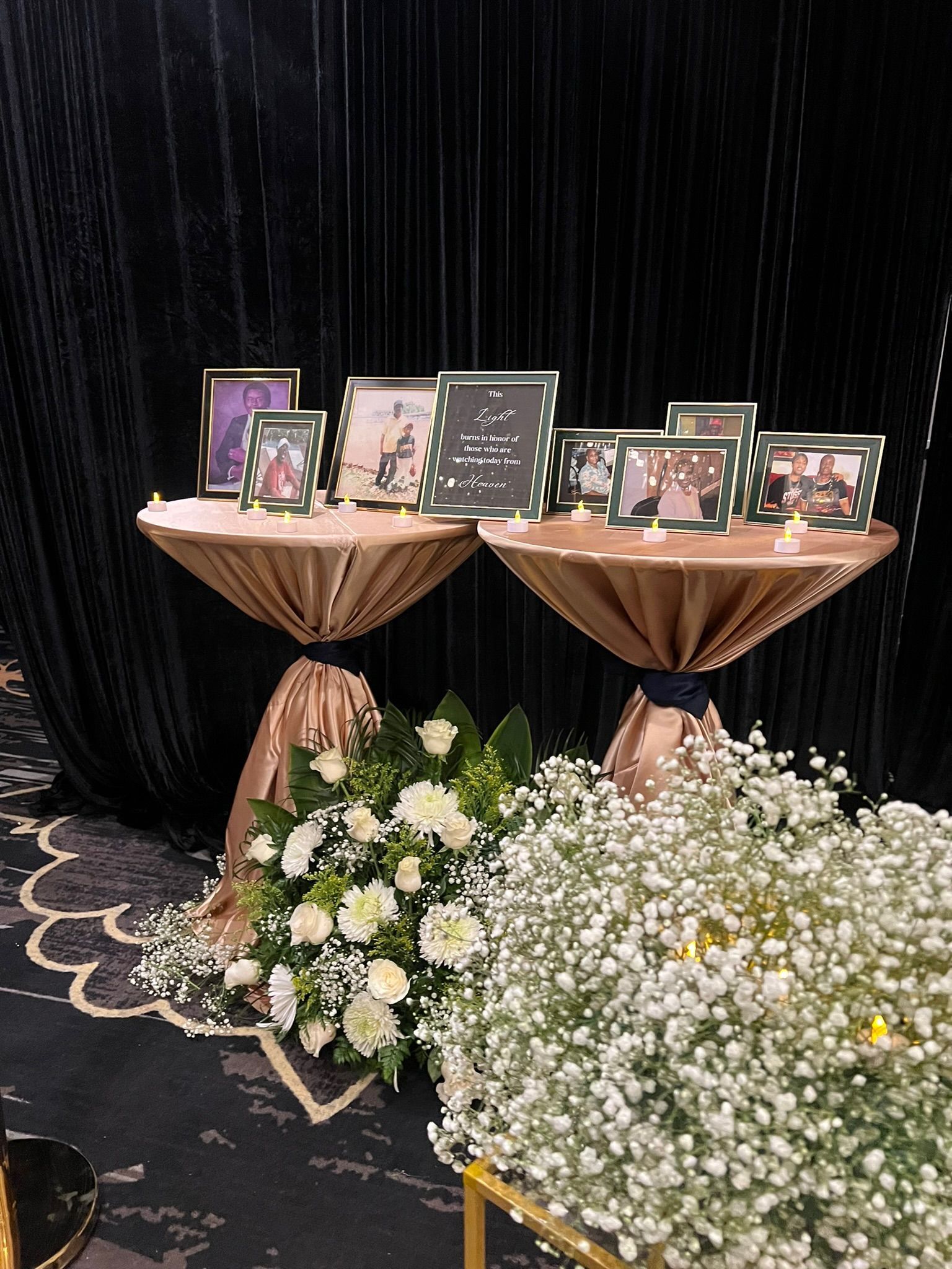 Two tables display framed photos, draped in gold fabric with floral arrangements in front of a dark backdrop.