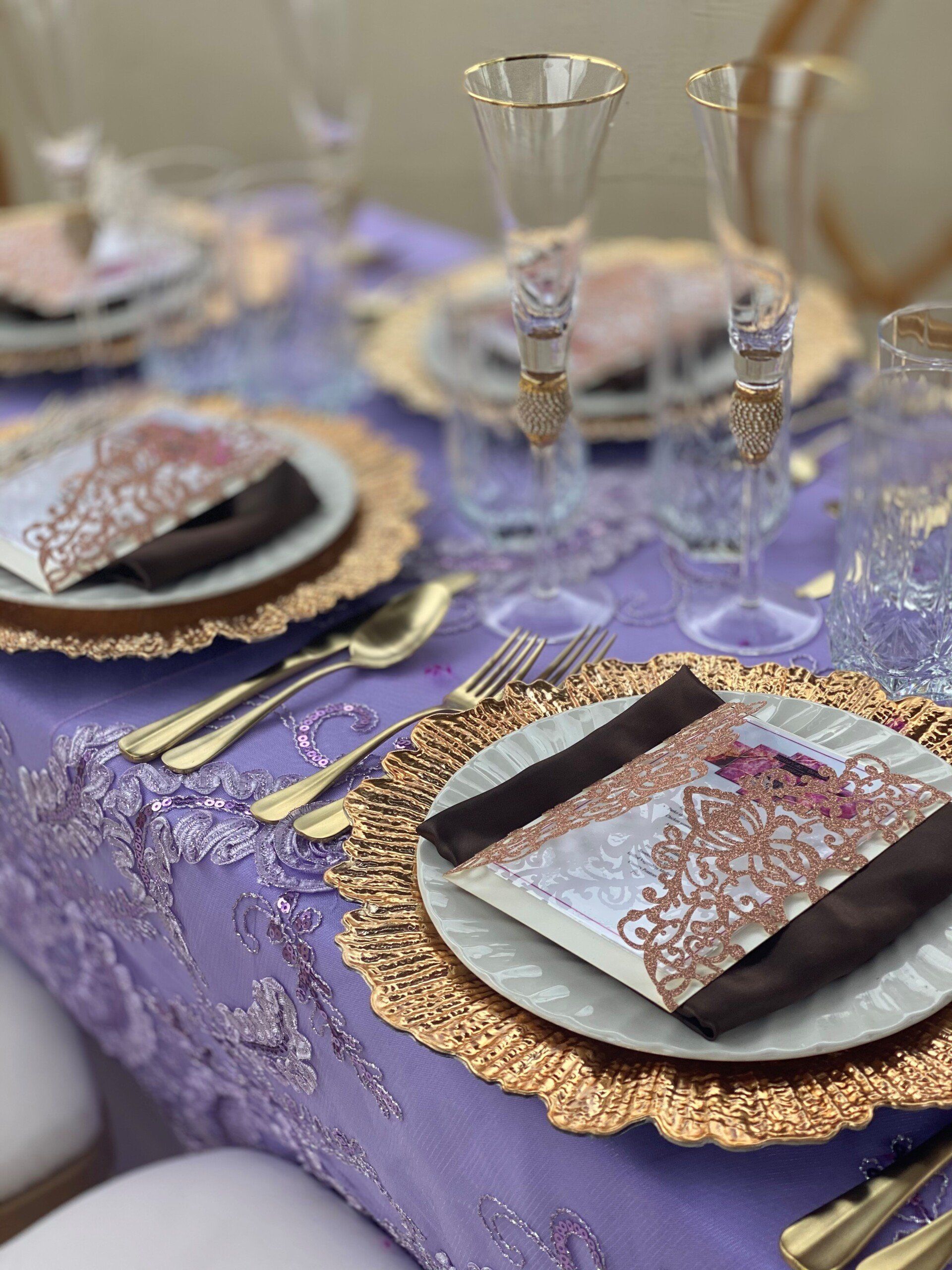 Elegant table setting with gold-rimmed plates, champagne flutes, gold silverware, and a purple lace tablecloth.