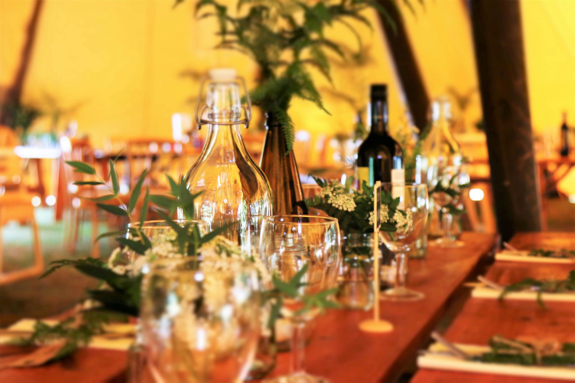 Table setting at an event with clear glasses, bottles, and greenery; inside a tent.