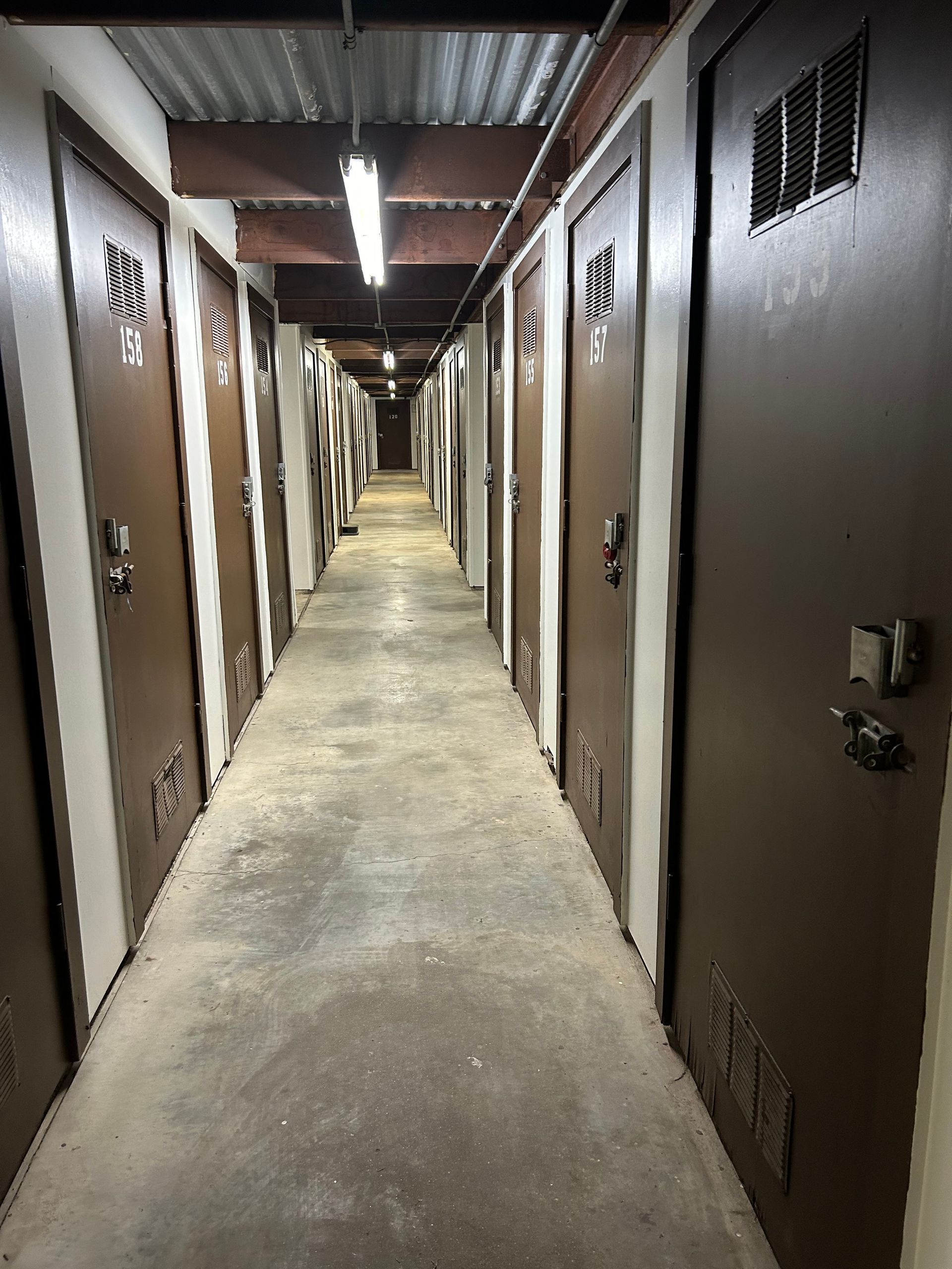 A long storage unit hallway with brown doors, concrete floor, and overhead lighting.