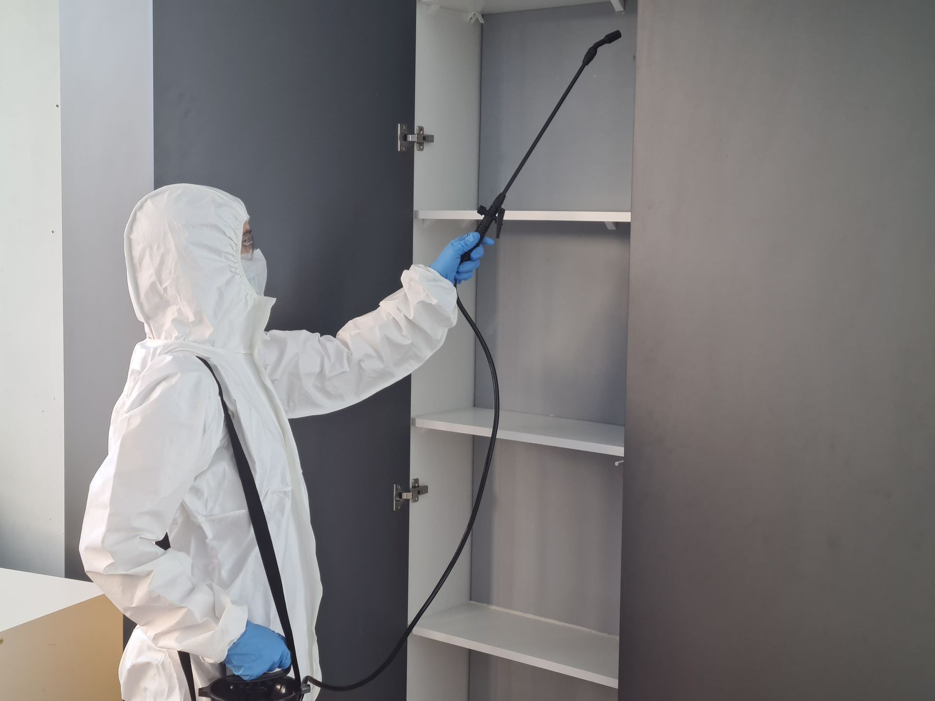 Person in white protective suit disinfecting a gray cabinet with a spray wand
