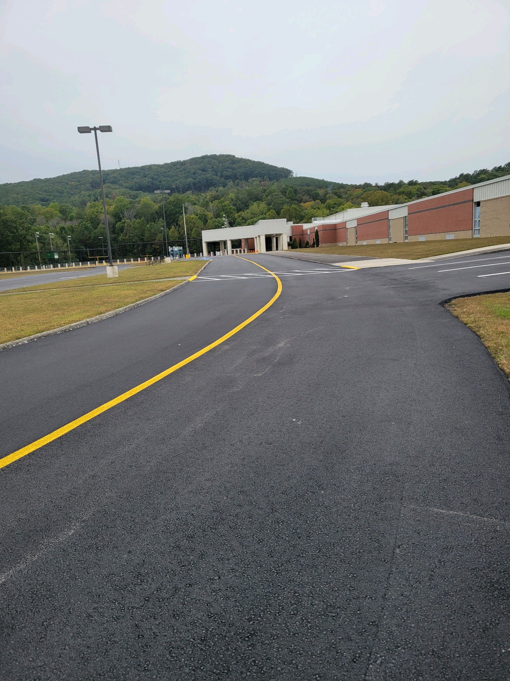 Asphalt road curves towards a brick building and a tree-covered hill on a cloudy day.