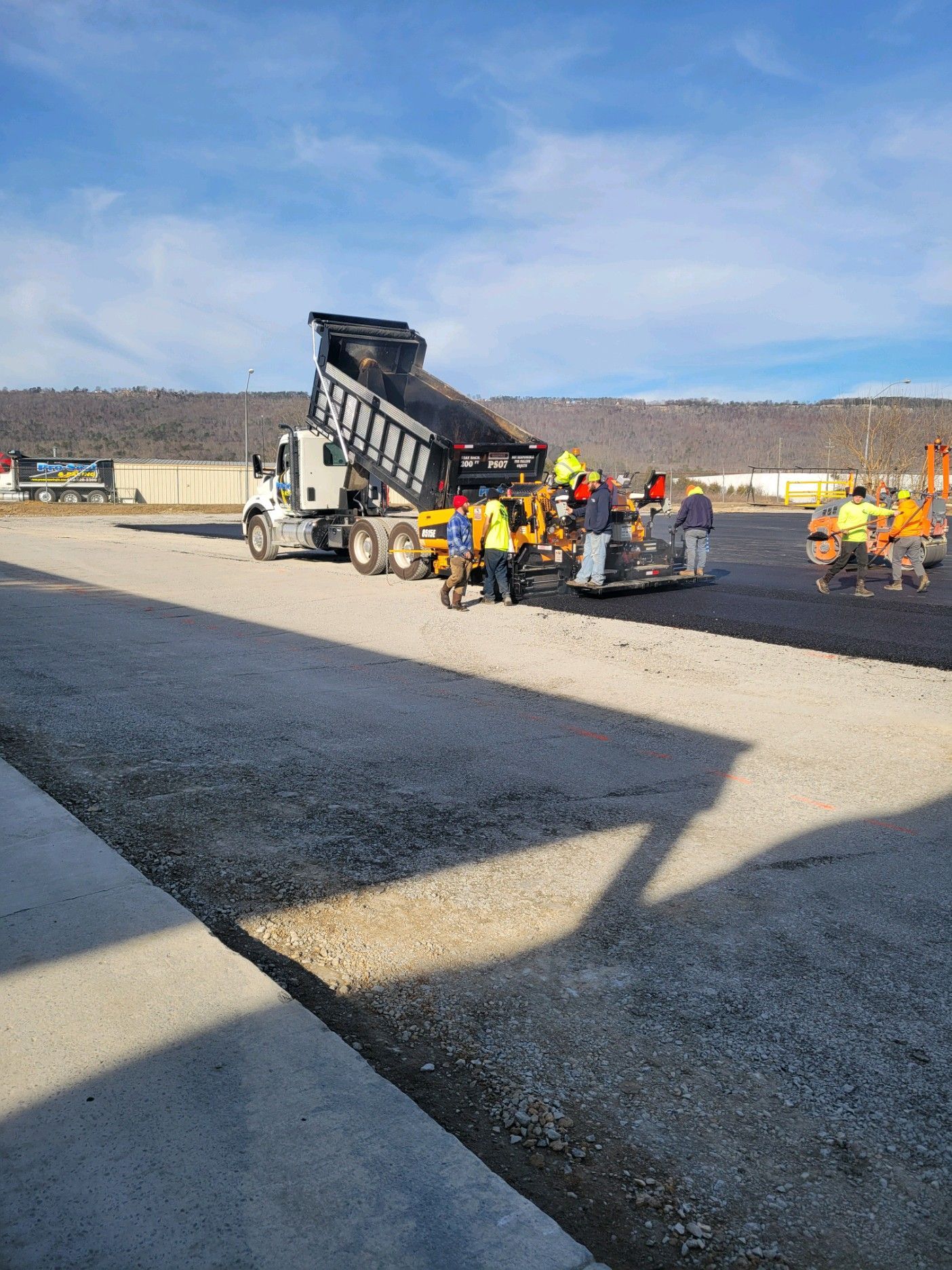 Dump truck unloading asphalt into a paving machine on a construction site. Workers in safety vests are present.