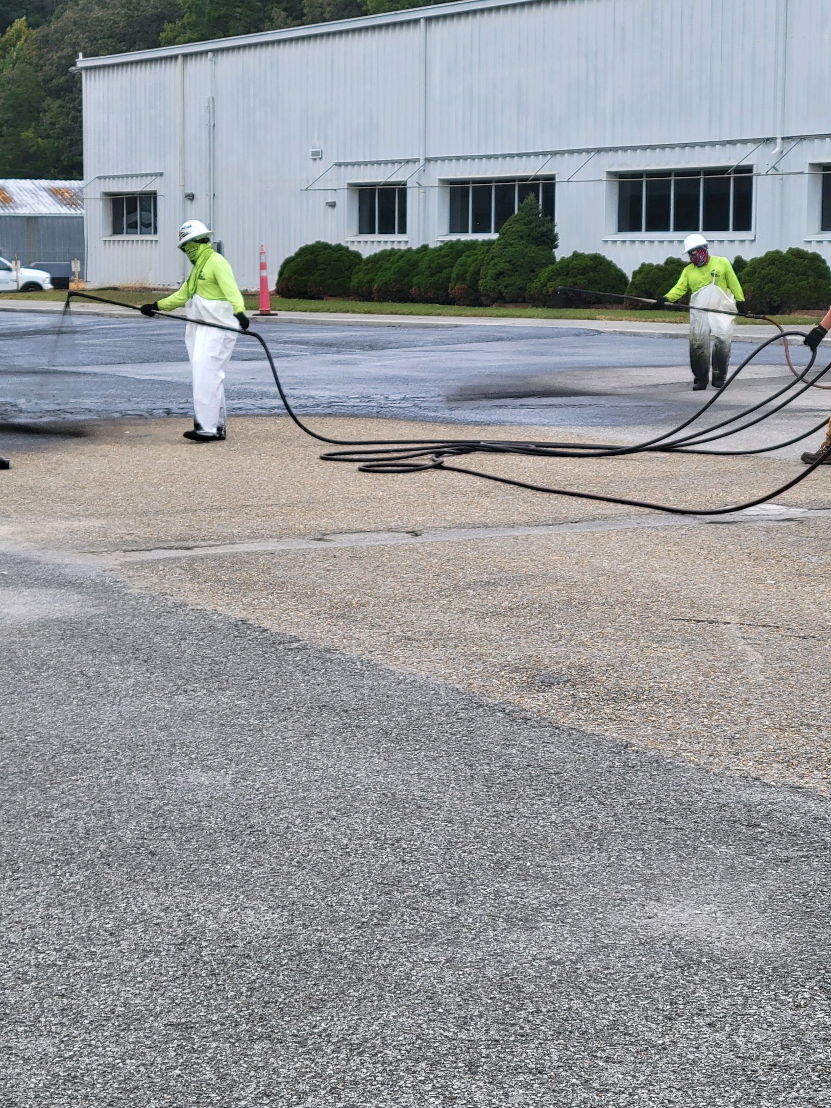 Two people in protective suits spray asphalt on a paved surface outside a building.