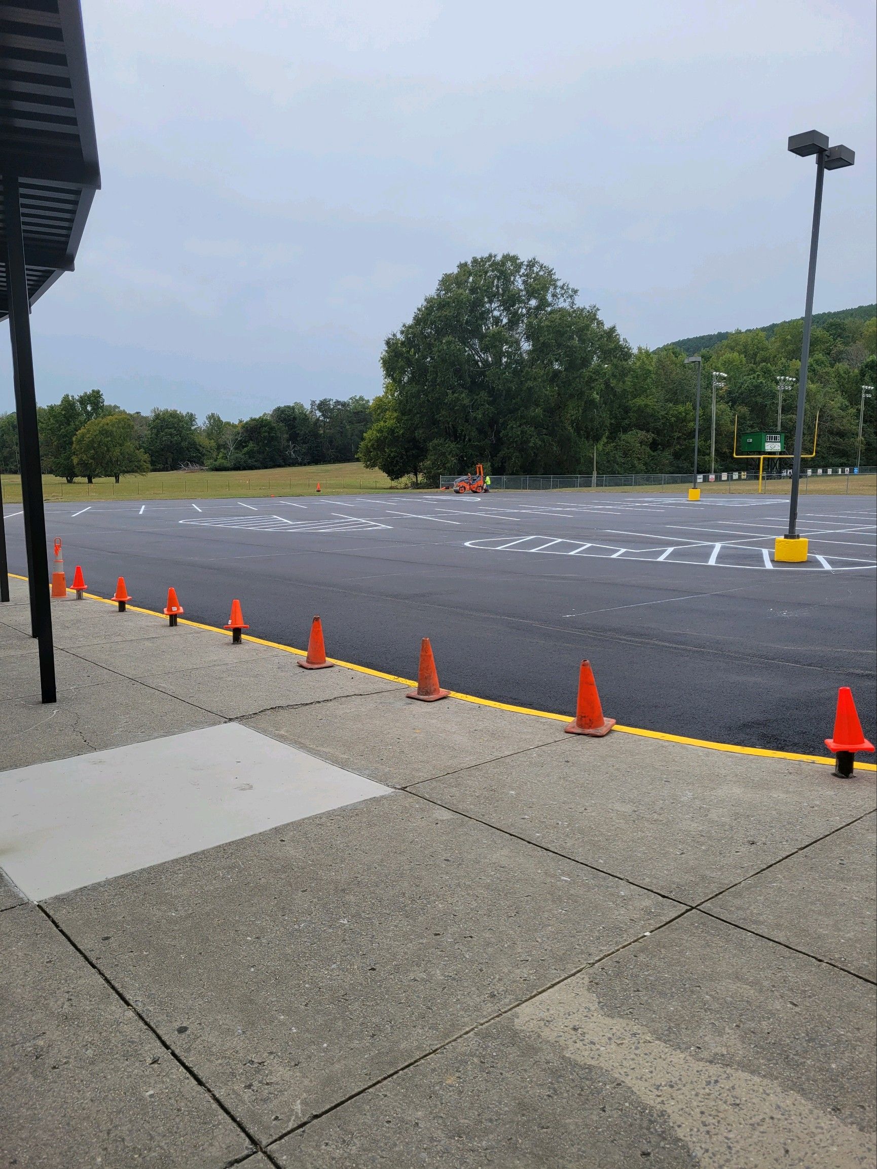 Orange cones line a parking lot's edge, marked spaces in asphalt, with trees and a cloudy sky in the background.