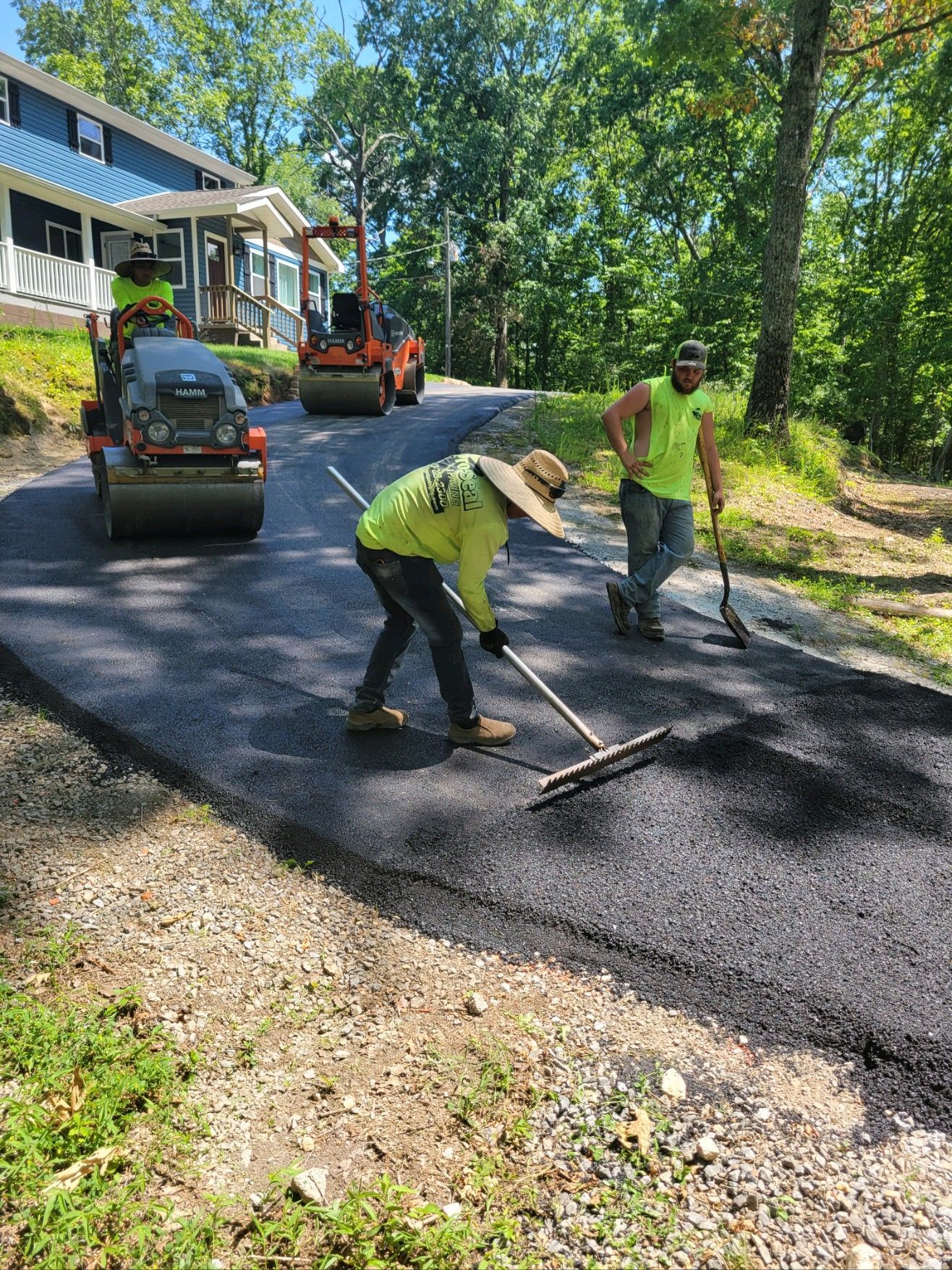 Asphalt paving of a driveway, with workers raking and two compactors rolling. Sunny outdoor setting.