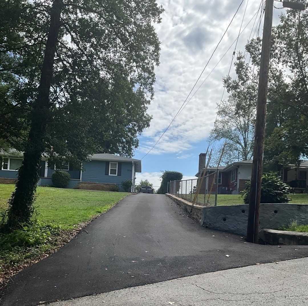 Asphalt driveway leading uphill between houses with trees and power lines, under a cloudy sky.
