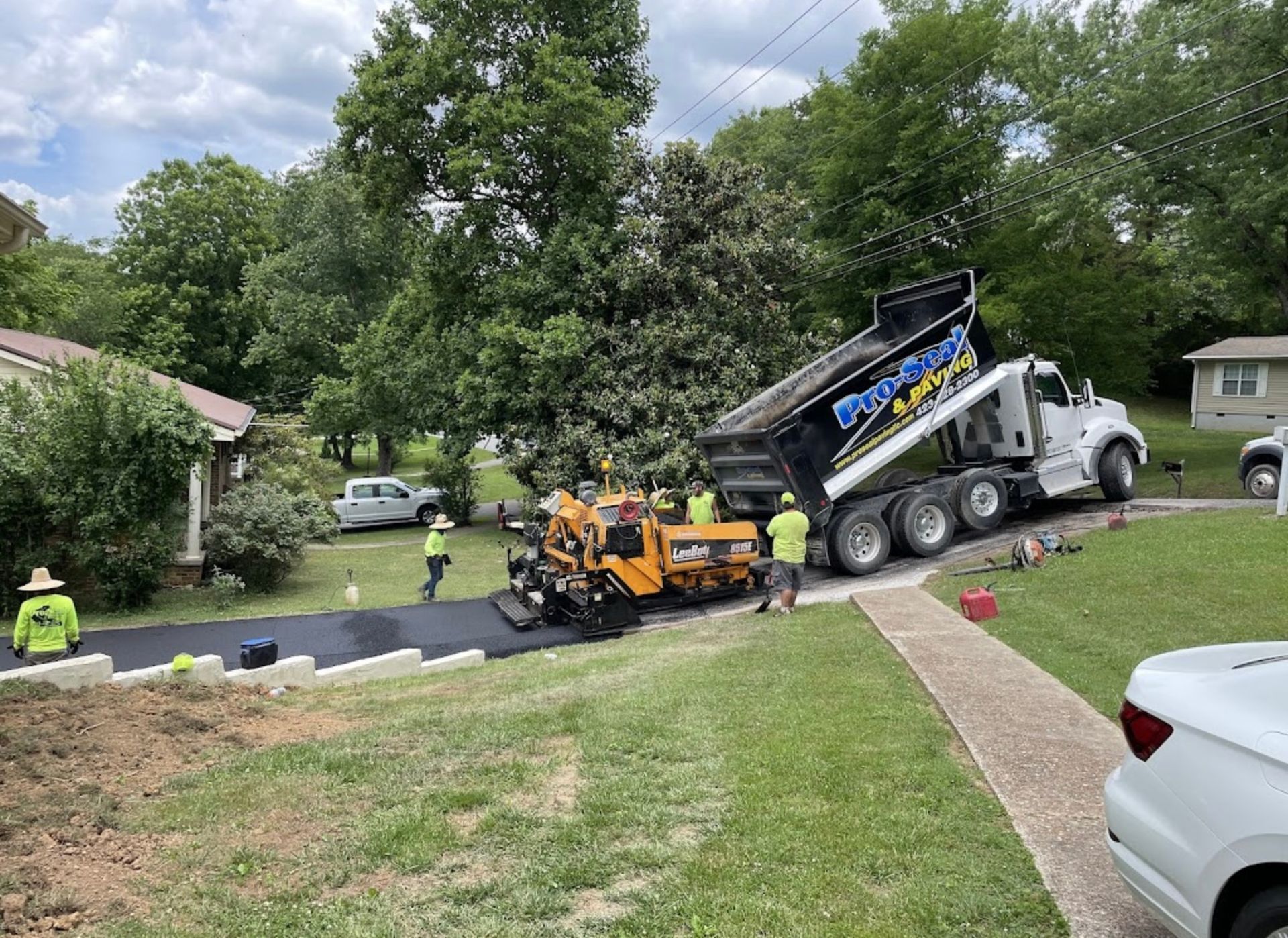 Asphalt paving crew at a residential driveway. Dump truck unloading into a paving machine. Workers in safety vests.