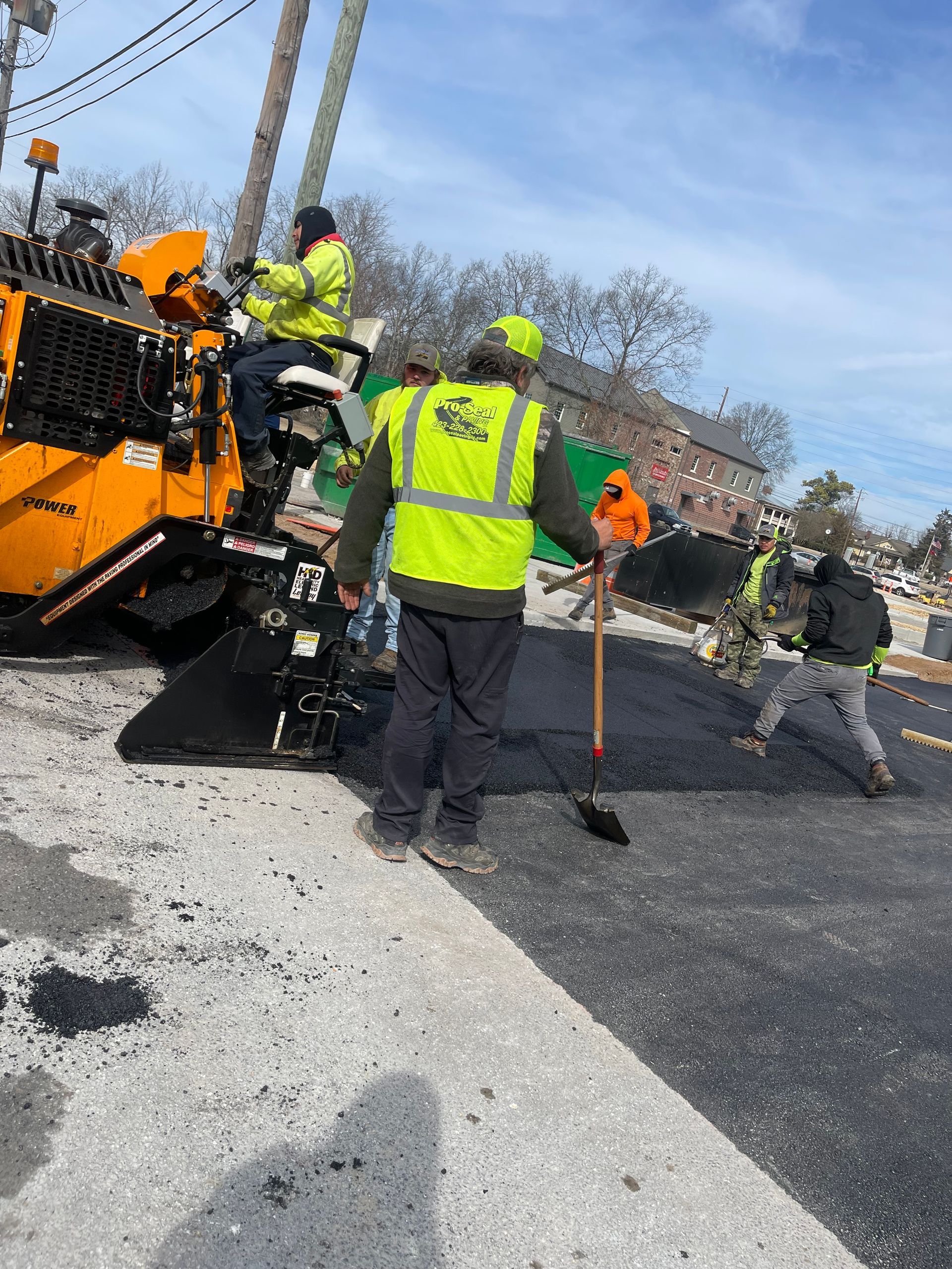 Road crew paving a street. Workers in safety vests use a paving machine and hand tools on a sunny day.